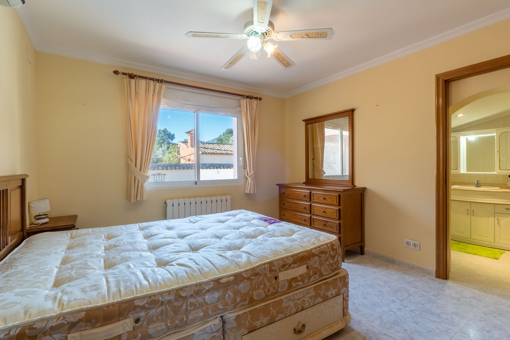 Hauptschlafzimmer mit Holzmöbeln, Deckenventilator und integriertem Bad. Der Raum ist mit einem Heizkörper und einem Fenster mit Außenblick ausgestattet.