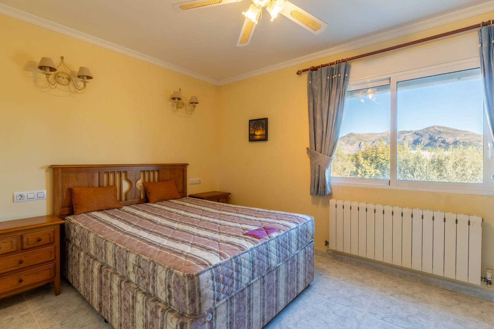 Helles Schlafzimmer mit klassischen Holzmöbeln, Deckenventilator und großem Fenster mit weitem Blick auf die Berge und die Natur von Jalón.