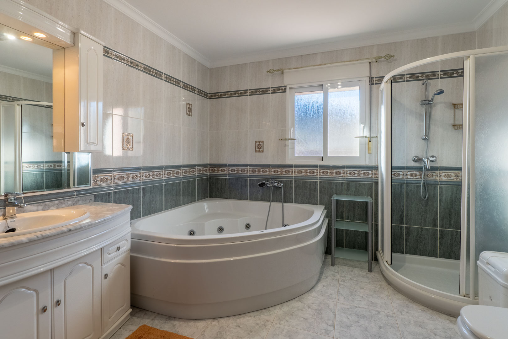 Komplettes Badezimmer mit Eck-Whirlpool-Badewanne, separater Runddusche und Waschtisch mit Marmorplatte und dekorativen Wandfliesen.