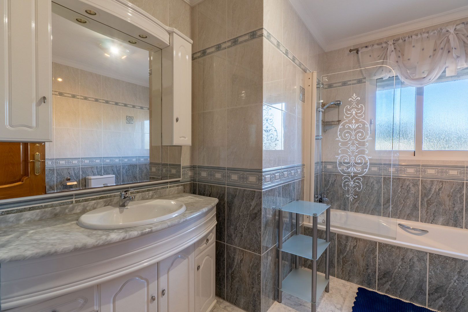 Komplettes Badezimmer mit Marmor-Waschtisch, weißen Schränken und Badewanne mit verzierter Glaswand. Geflieste Wände mit Zierbordüren und Außenfenster.
