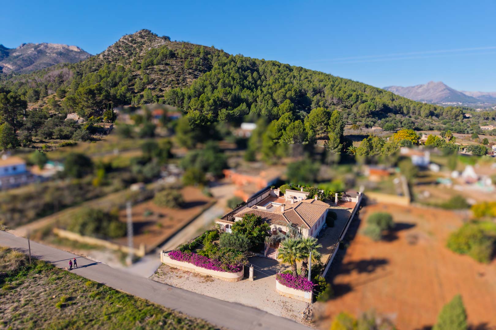 Luftbild der mediterranen Villa auf ebenem Grundstück mit traditionellem Ziegeldach und Bougainvillea-Gärten vor der Kulisse der umliegenden Berge.