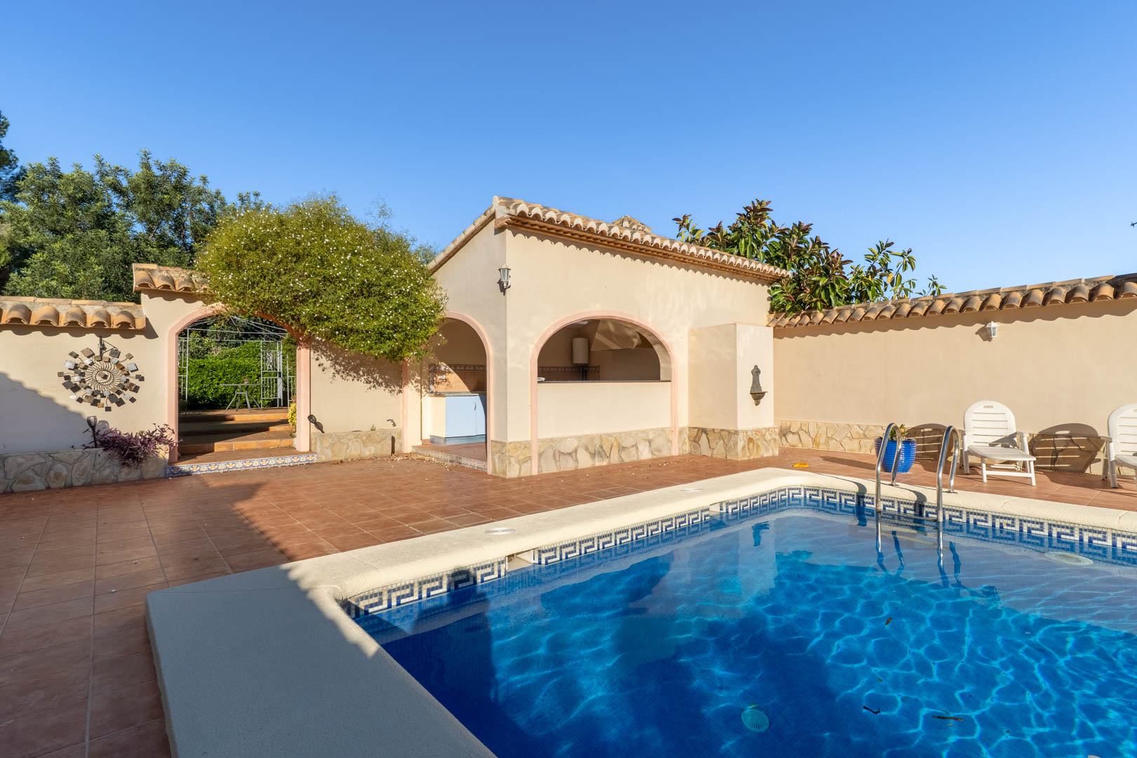 Poolbereich mit Terrakotta-Terrasse, überdachter Außenküche mit Rundbögen und Natursteinsockel in sonniger, privater Atmosphäre.