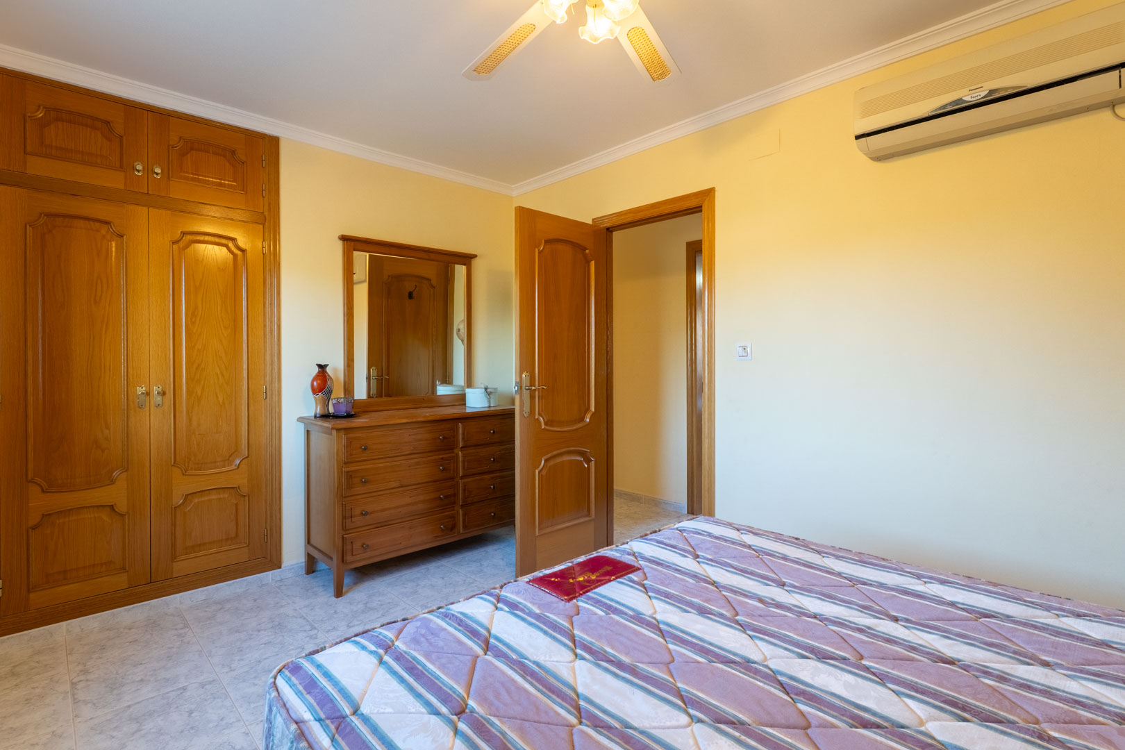 Schlafzimmer mit Einbauschränken aus Holz, passender Kommode mit Spiegel, Klimaanlage und Deckenventilator auf hellen Fliesenböden.