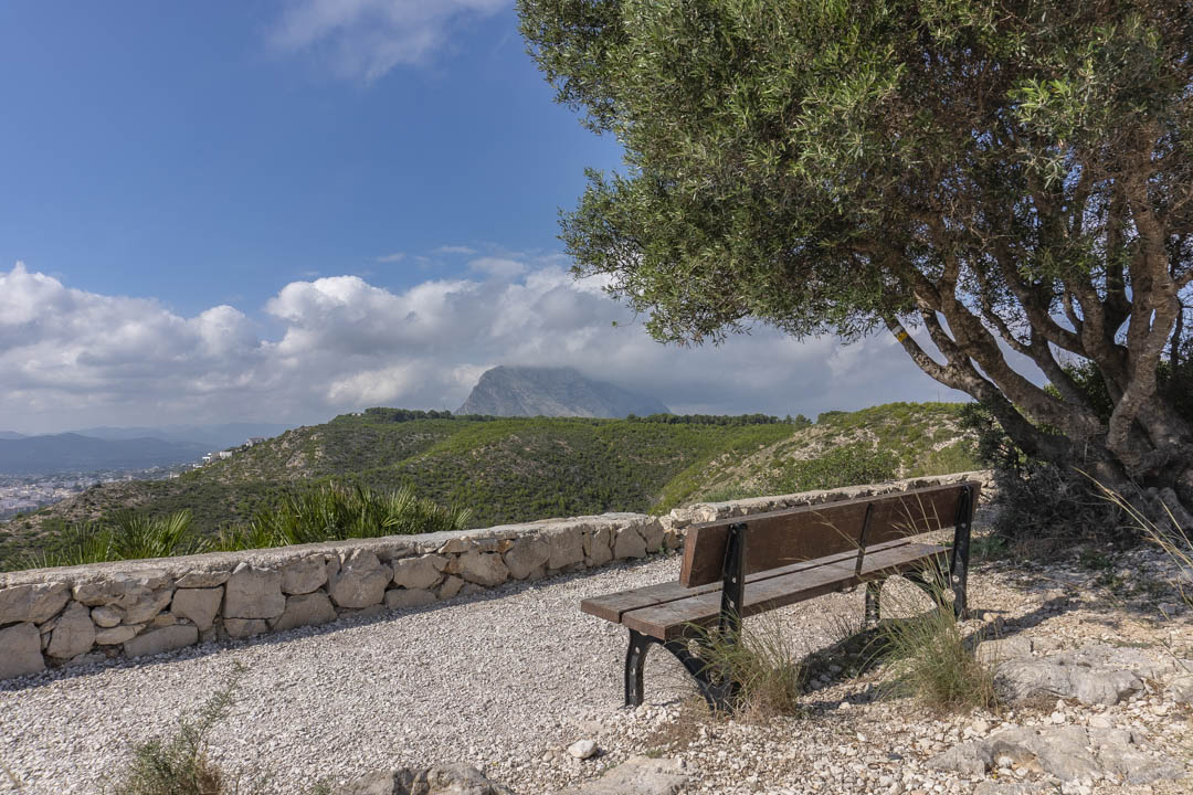 Aussichtspunkt mit Holzbank und Natursteinmauer, der einen weiten Blick auf das Montgó-Massiv und die umliegende Hügellandschaft bietet.