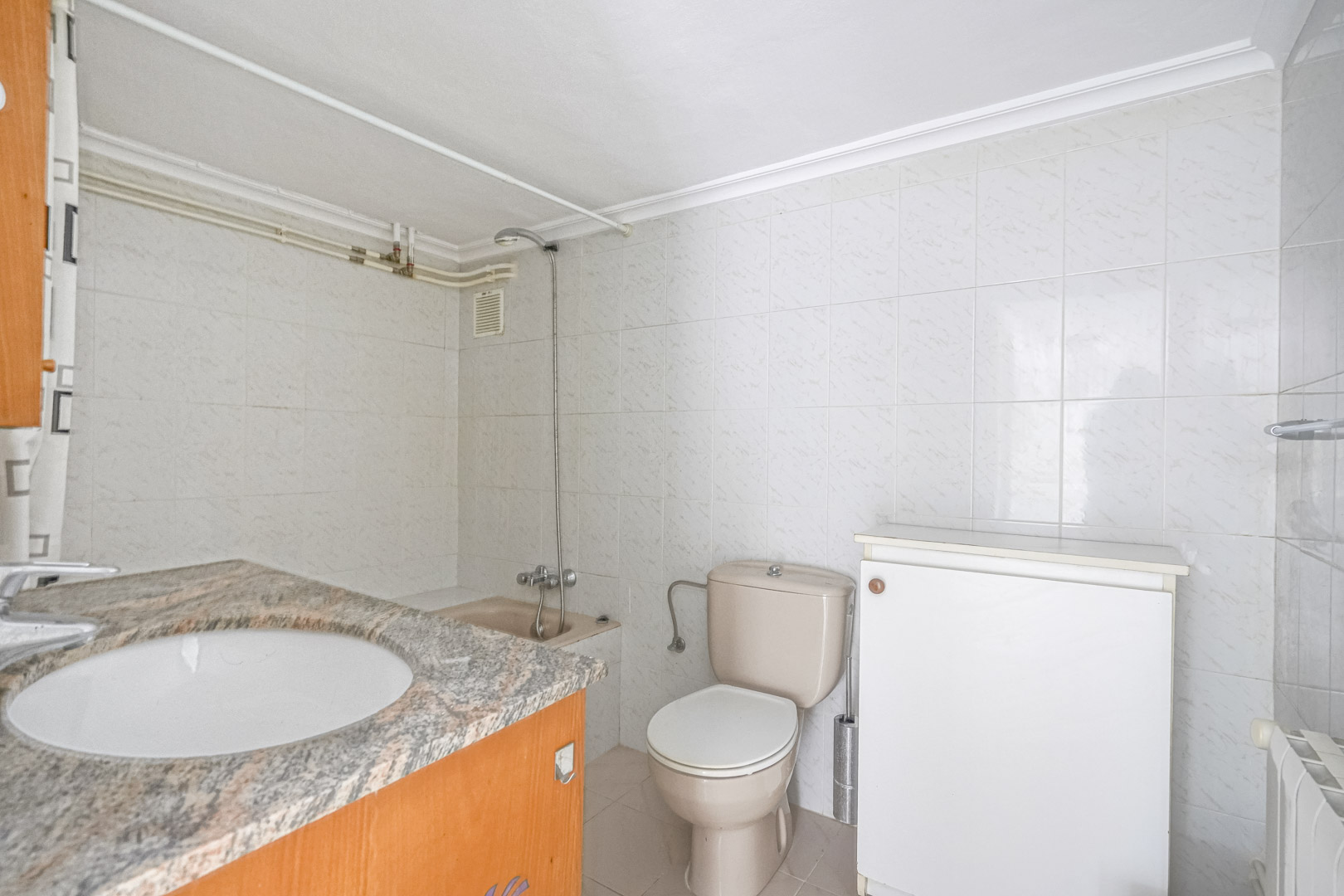 Badezimmer mit Granit-Arbeitsplatte, Einbauspülbecken, beigefarbener Toilette und weiß gefliesten Wänden. Ausgestattet mit Duschbereich und Heizkörper.