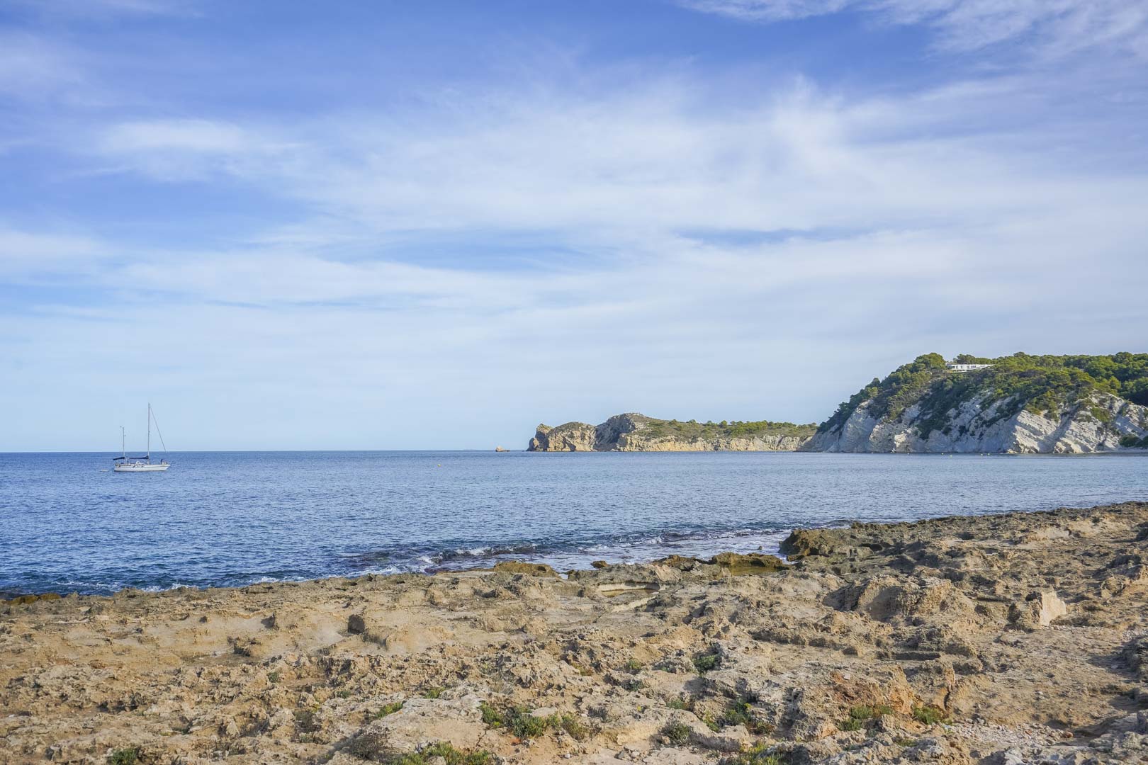 Blick auf die felsige Küste von Jávea mit dem Mittelmeer, Kalksteinklippen und einem Segelboot unter klarem blauem Himmel.
