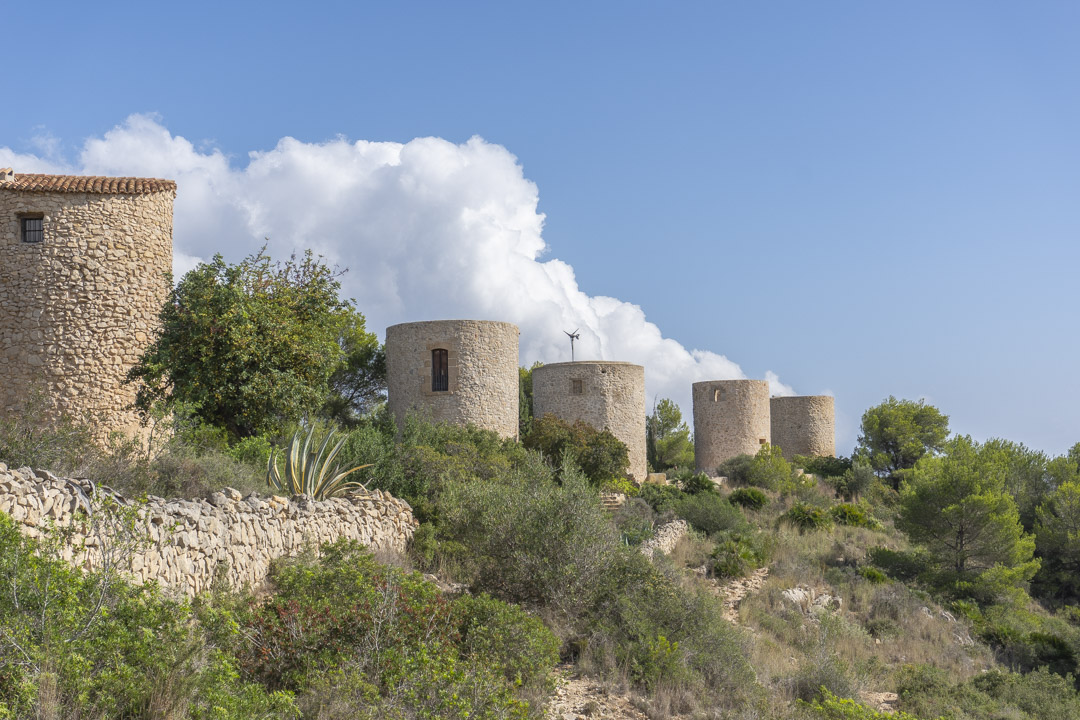 Blick auf die historischen Steinwindmühlen von Jávea auf einem Hügel mit mediterraner Vegetation, Trockenmauern und klarem blauem Himmel.