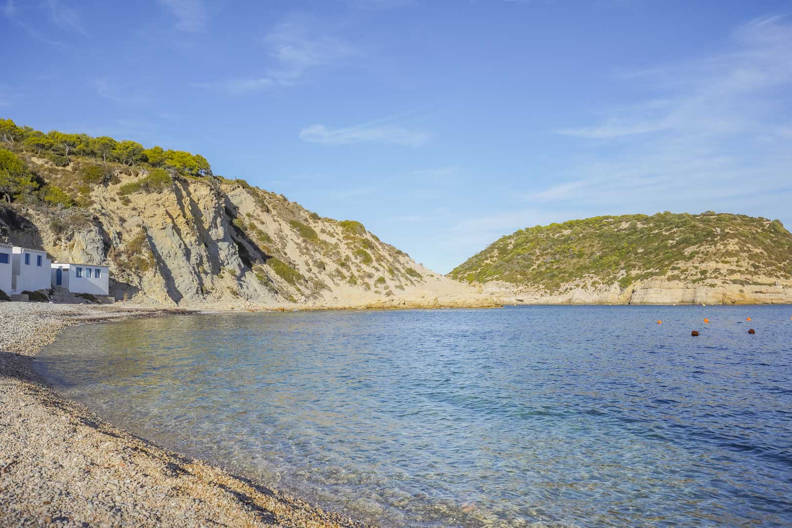 Blick auf eine Kieselbucht in Jávea mit kristallklarem Wasser, umgeben von felsigen Klippen und mediterraner Vegetation unter blauem Himmel.