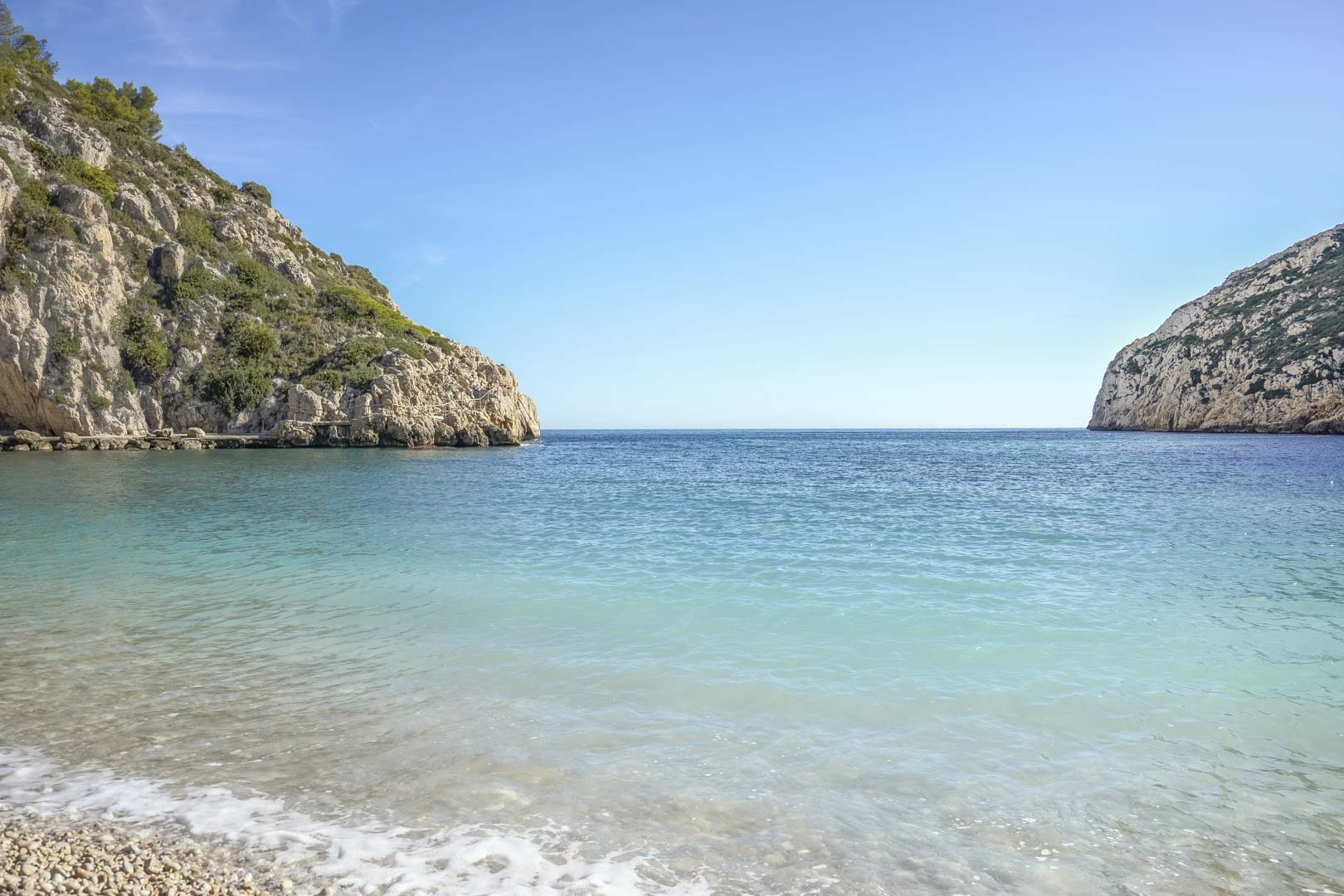 Blick auf eine mediterrane Bucht mit türkisblauem Wasser und Kieselstrand, umgeben von markanten Felsklippen in der Nähe des Standorts in Jávea.