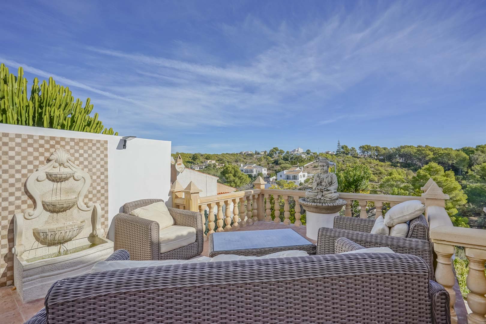 Erhöhte Terrasse mit Rattan-Sitzgruppe, dekorativem Wandbrunnen und Steinbalustrade mit weitem Blick über die Hügellandschaft von Jávea.