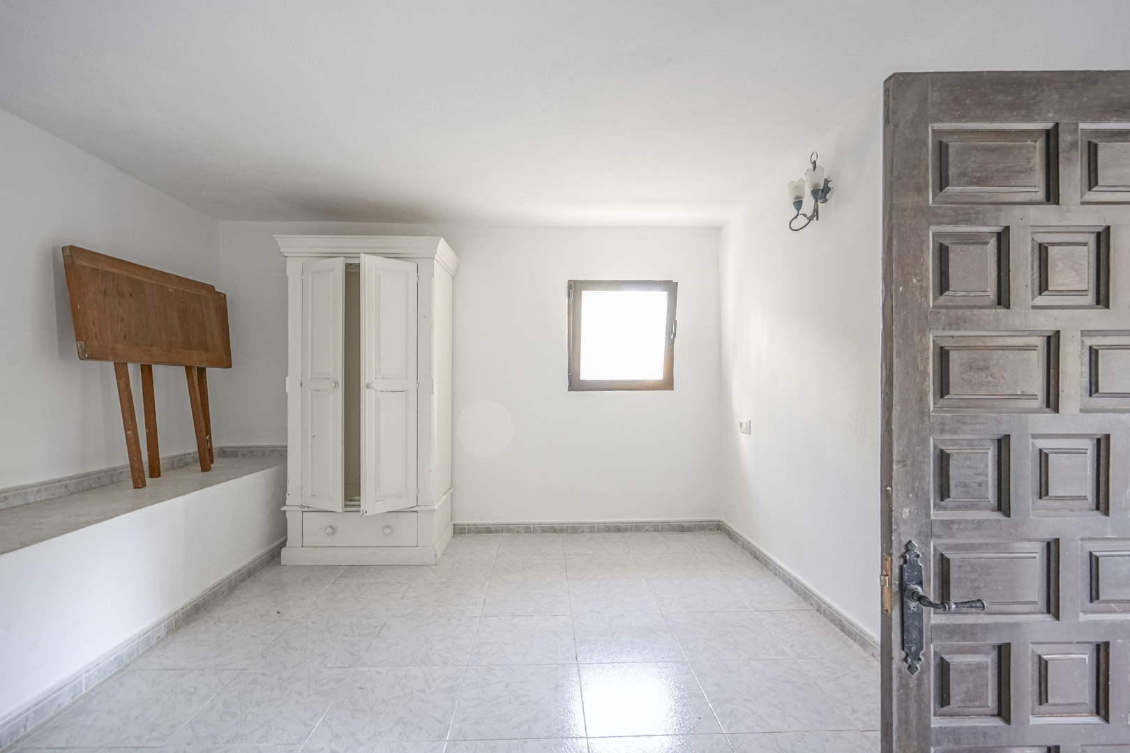 Heller Raum mit hellen Fliesenböden, weißen Wänden und einer traditionell geschnitzten Holztür. Ein freistehender weißer Schrank und ein Fenster sorgen für Licht.