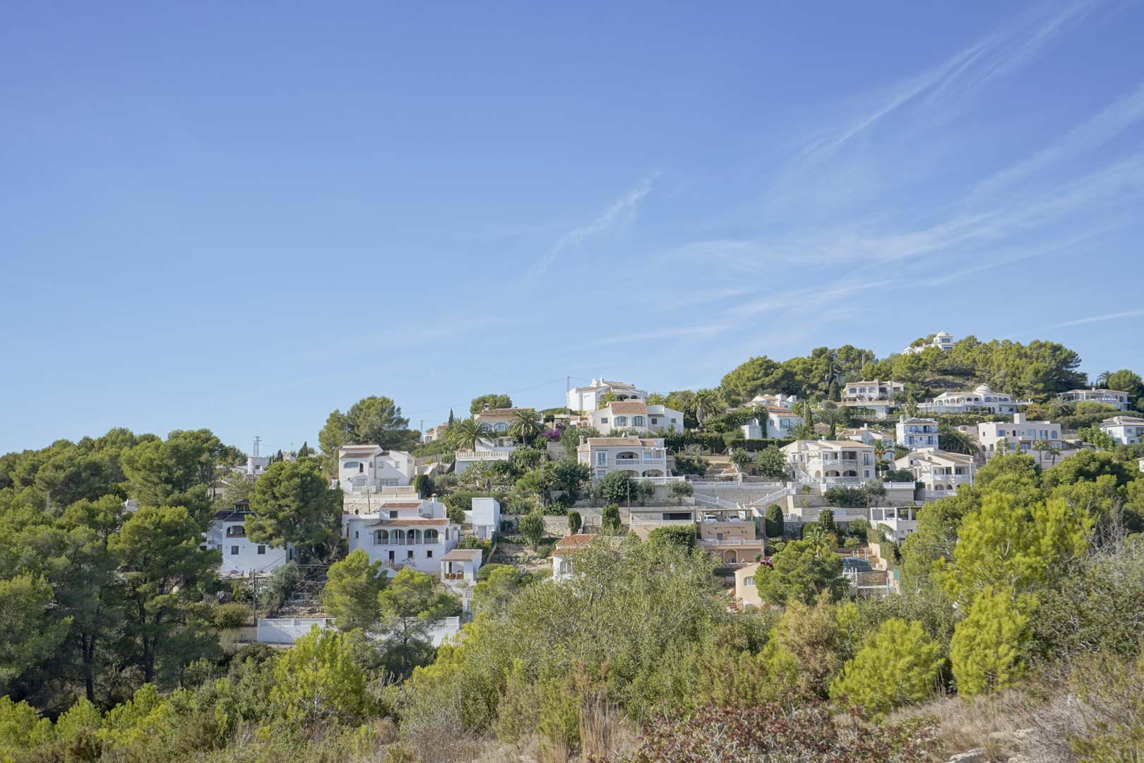 Panoramablick auf das Wohngebiet Pinomar in Jávea mit traditionellen weißen Villen, die in einen bewaldeten Hang unter blauem Himmel eingebettet sind.