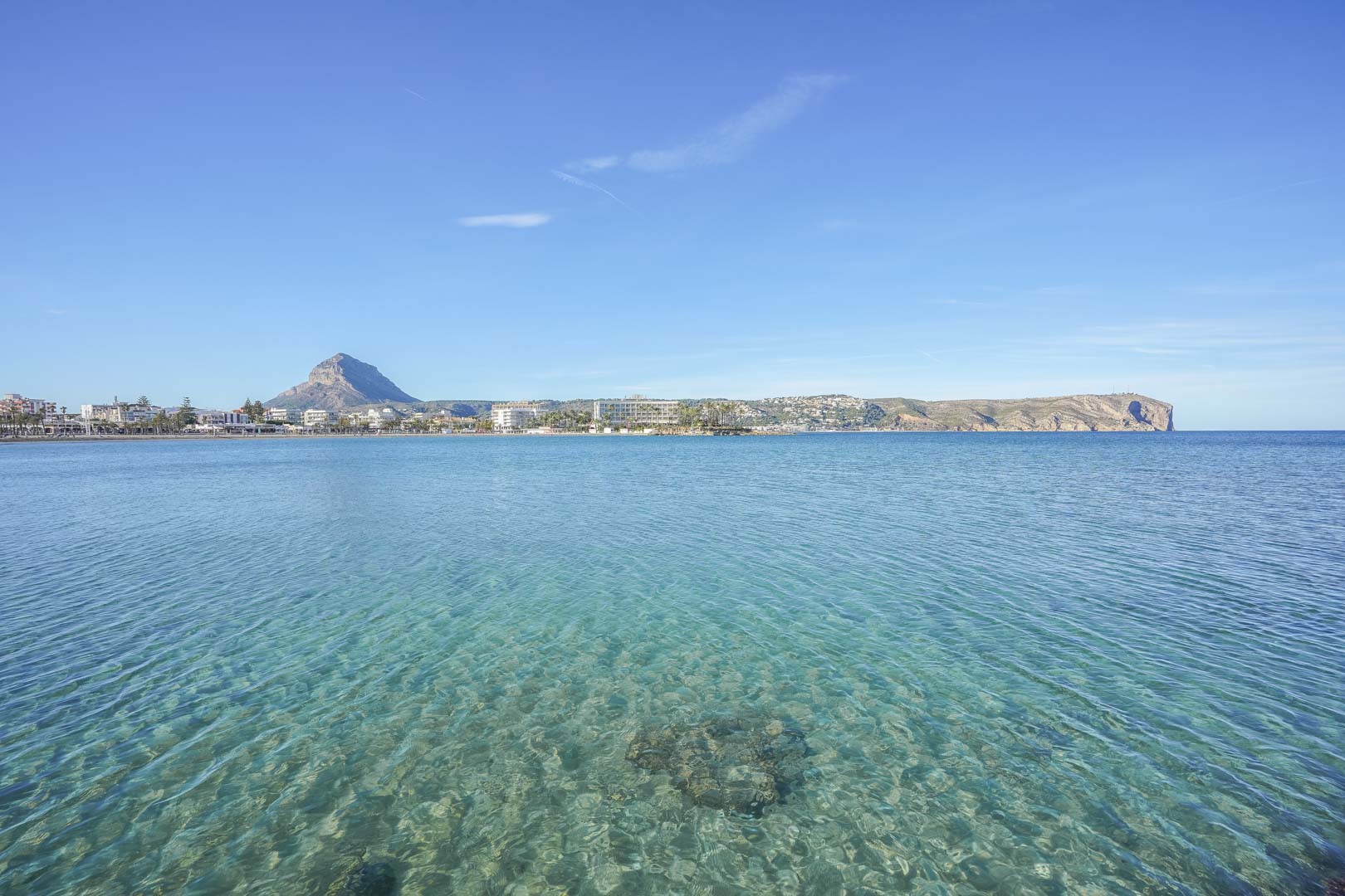 Panoramablick auf die Bucht von Jávea mit kristallklarem, türkisfarbenem Wasser, dem Berg Montgó im Hintergrund und der Küstenlinie unter blauem Himmel.