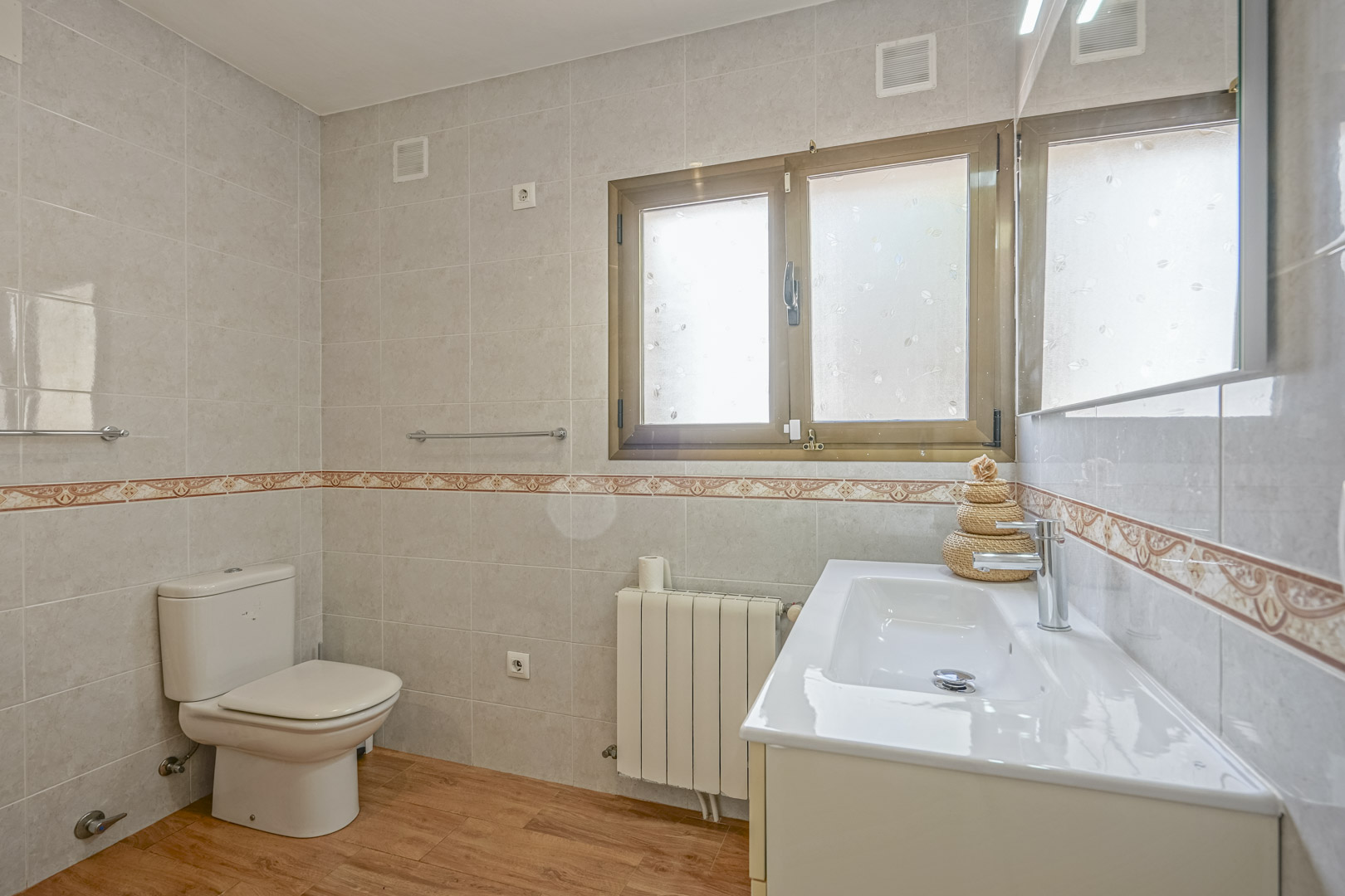 Renoviertes Badezimmer mit Boden in Holzoptik, gefliesten Wänden mit Zierborte, weißem Waschtisch und Heizkörper unter dem Milchglasfenster.