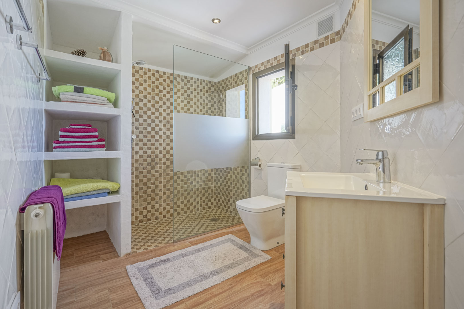Renoviertes Badezimmer mit ebenerdiger Mosaikdusche, gemauerten Einbauregalen, Boden in Holzoptik und einem Waschtisch aus hellem Holz.