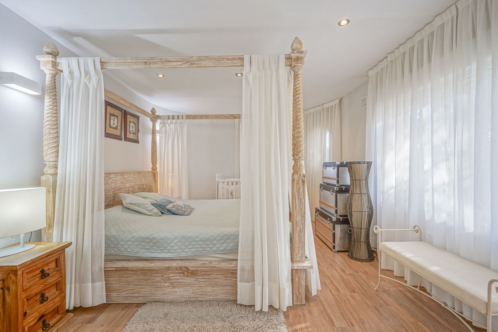 Schlafzimmer mit einem geschnitzten Himmelbett aus Holz, Laminatboden und hellen Fenstern mit lichtdurchlässigen weißen Vorhängen.