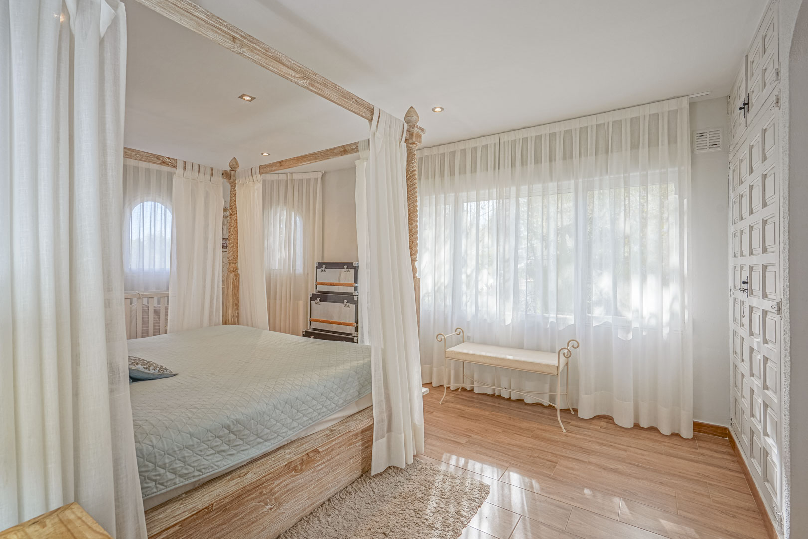 Schlafzimmer mit einem Himmelbett aus hellem Holz, Keramikboden in Holzoptik und großen Fenstern mit transparenten Vorhängen für sanftes Tageslicht.