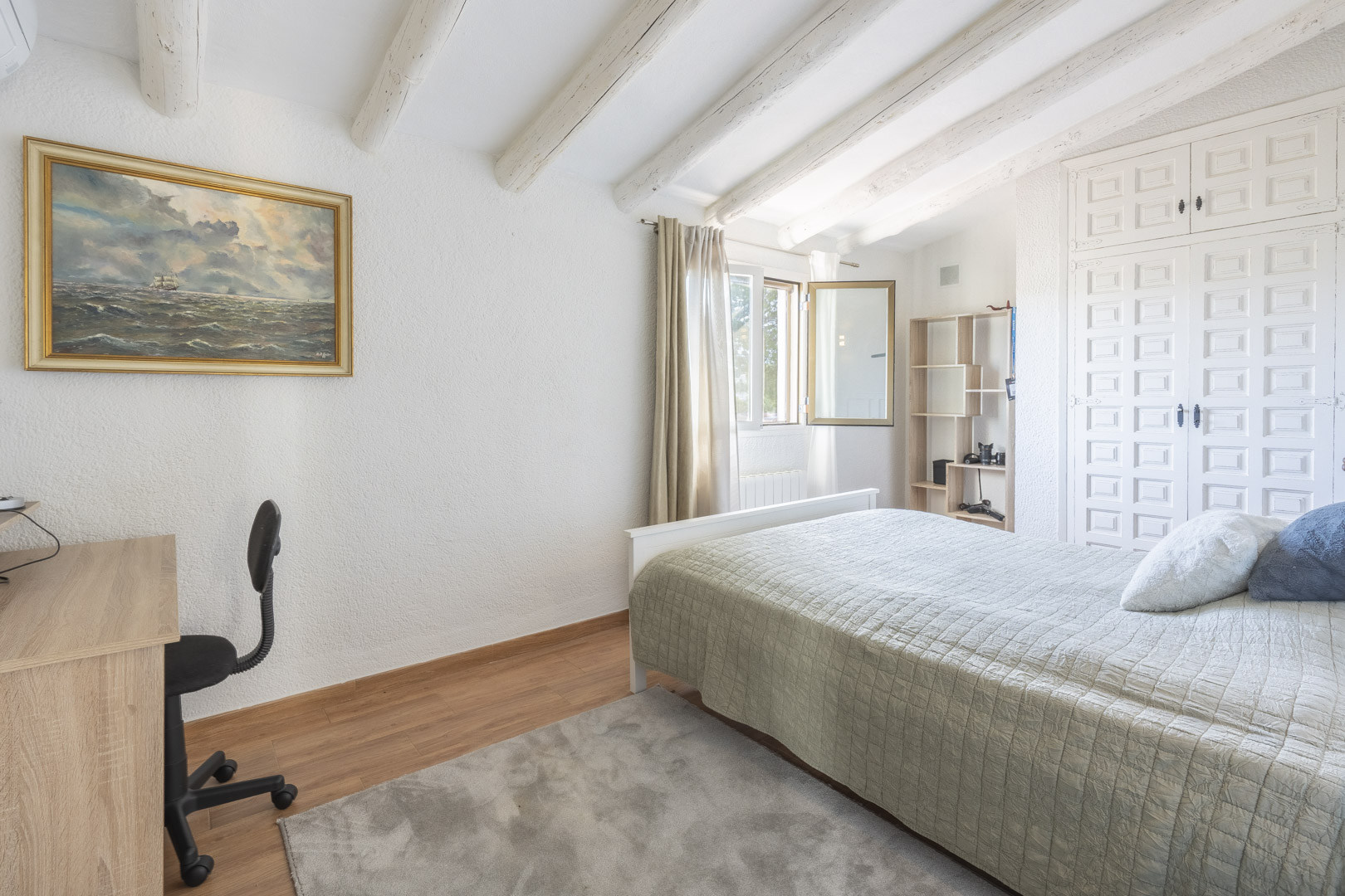 Schlafzimmer mit weiß gestrichenen Holzbalken, Dachschrägen, traditionellen Einbauschränken und einem hellen Arbeitsbereich am Fenster.