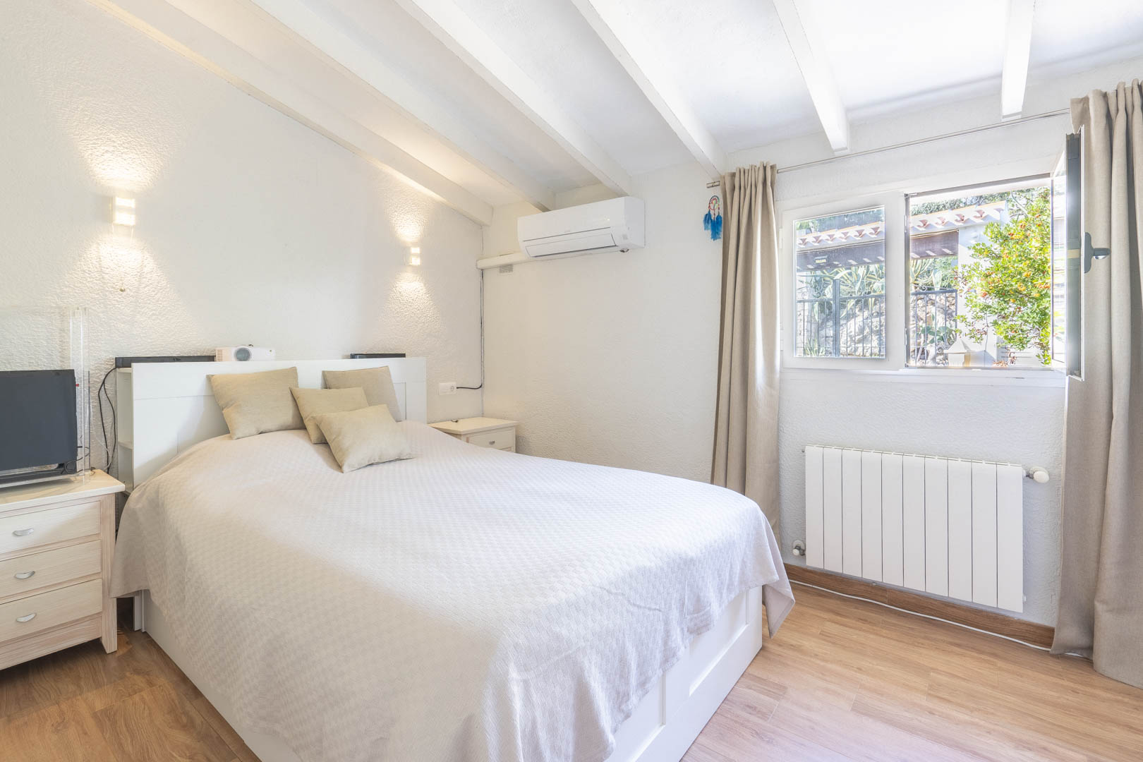 Schlafzimmer mit weißer Balkendecke, Klimaanlage und Heizkörper. Der Raum verfügt über Laminatboden und ein Fenster mit Tageslicht.