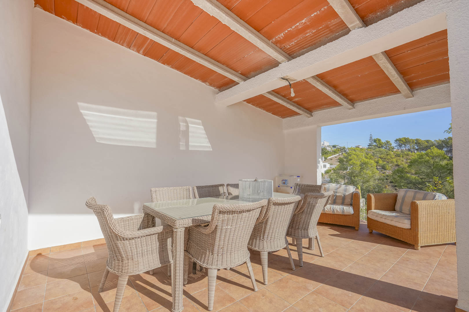 Überdachte Terrasse mit Balkendecke und Terrakotta-Elementen, ausgestattet mit einer Korb-Essgruppe und freiem Blick auf die umliegende Pinienlandschaft.
