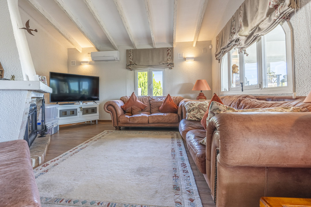 Wohnzimmer mit weißen Deckenbalken, Ledersofas, einem gemauerten Kamin und moderner Split-Klimaanlage an der Wand.