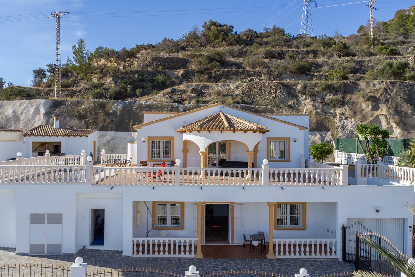 Außenansicht der zweistöckigen Villa mit weißen Fassaden, klassischen Balustraden und überdachten Veranden vor der Kulisse der Sierra de Oltà.