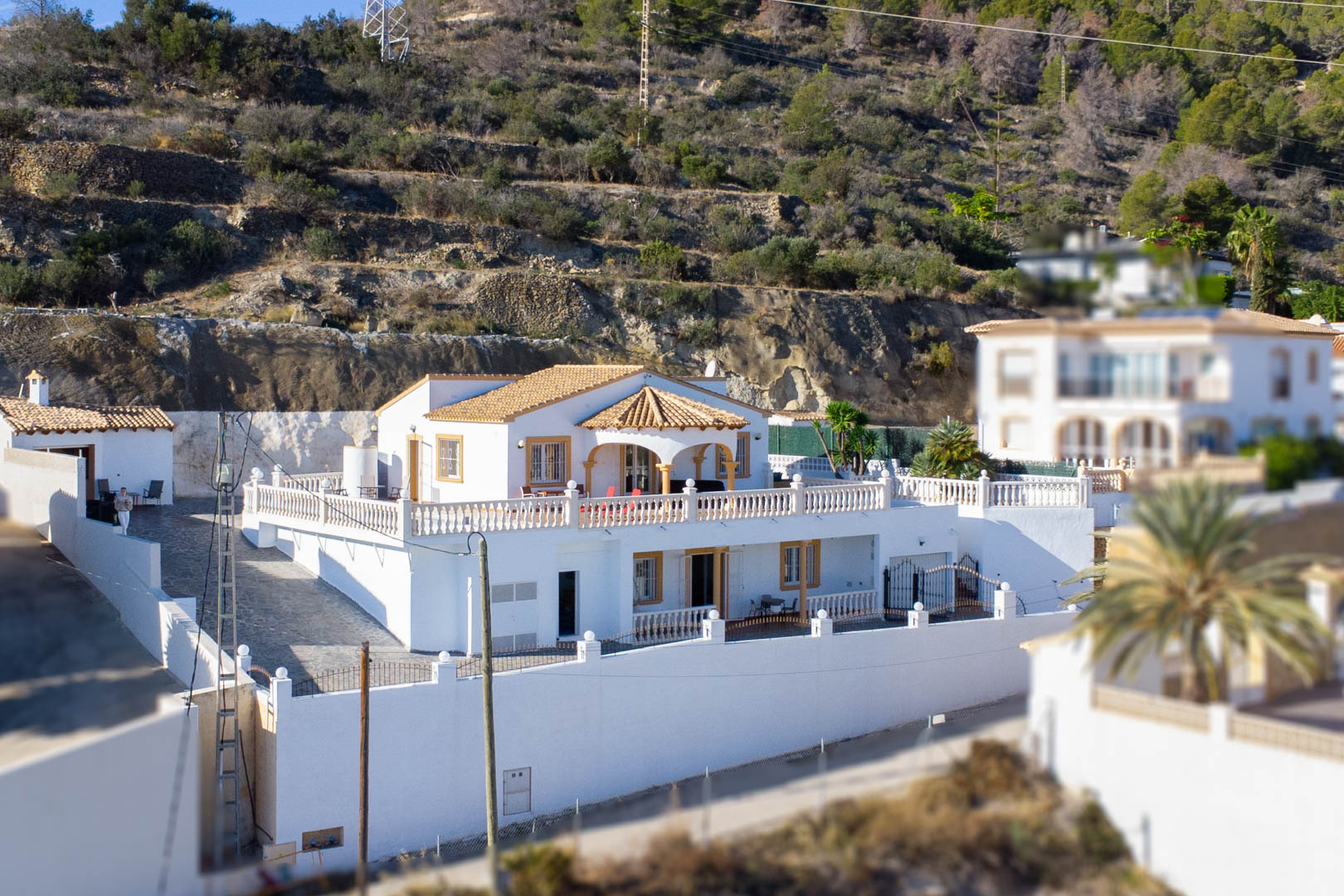 Außenansicht der zweistöckigen Villa mit weißer Fassade, Terrakottadach und weitläufigen Terrassen mit Steinbalustraden am Hang der Sierra de Oltà.