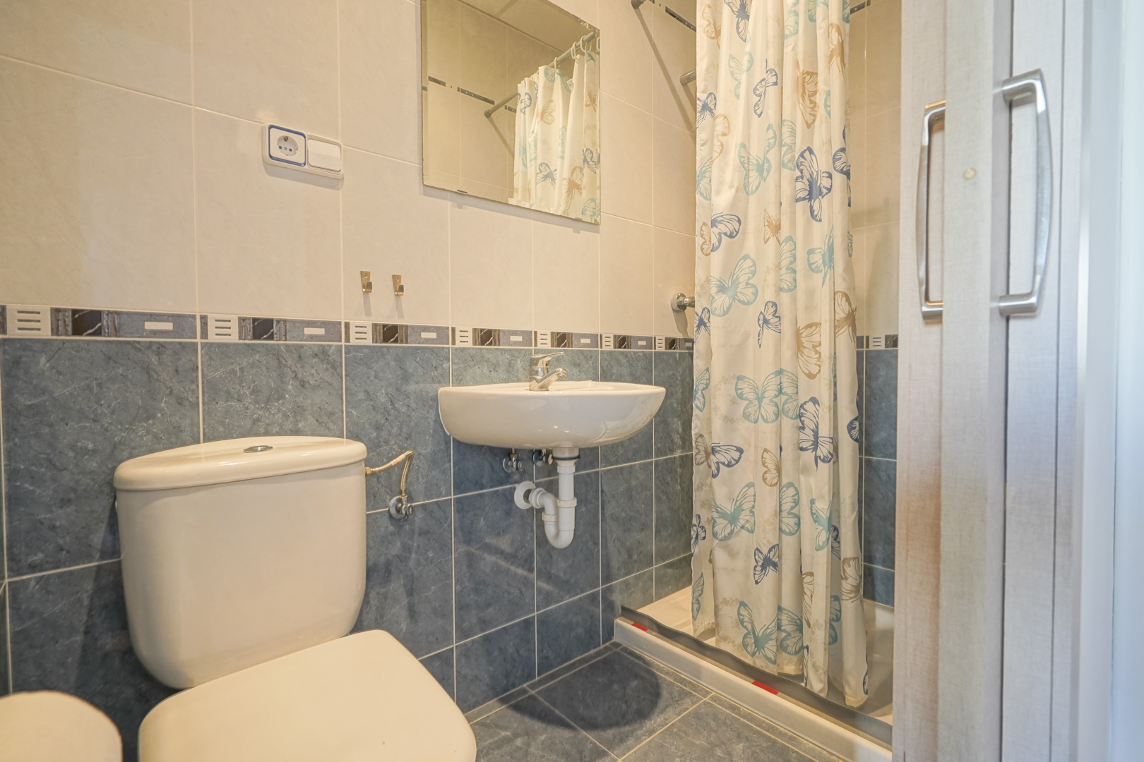 Badezimmer mit blau-beigen Fliesen, ausgestattet mit einem wandhängenden Waschbecken, WC und einem Duschbereich mit Schmetterlingsvorhang.