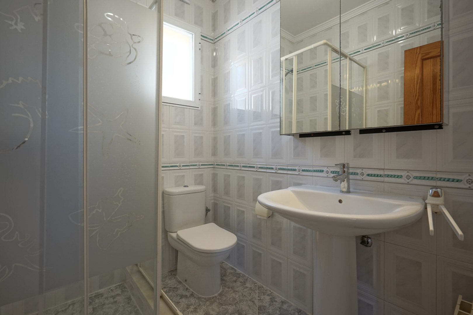 Badezimmer mit Standwaschbecken, WC und Dusche mit dekorierter Glaswand. Die Wände sind hell gefliest und verfügen über eine ornamentale Bordüre.