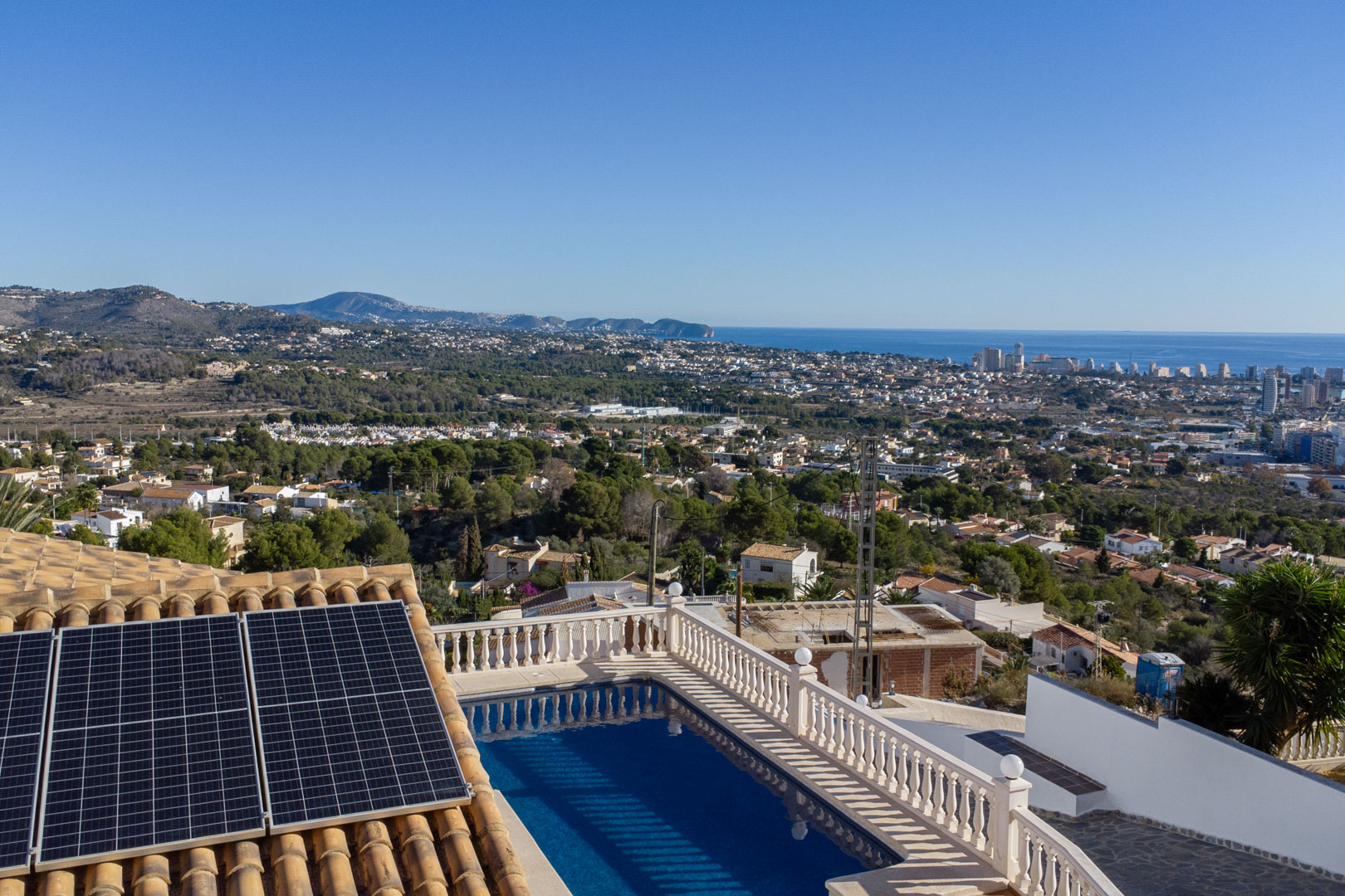 Erhöhter Blick auf den Pool mit Balustrade, Photovoltaik-Paneele auf dem Ziegeldach und das Panorama der Küste von Calpe mit Blick auf das Mittelmeer.