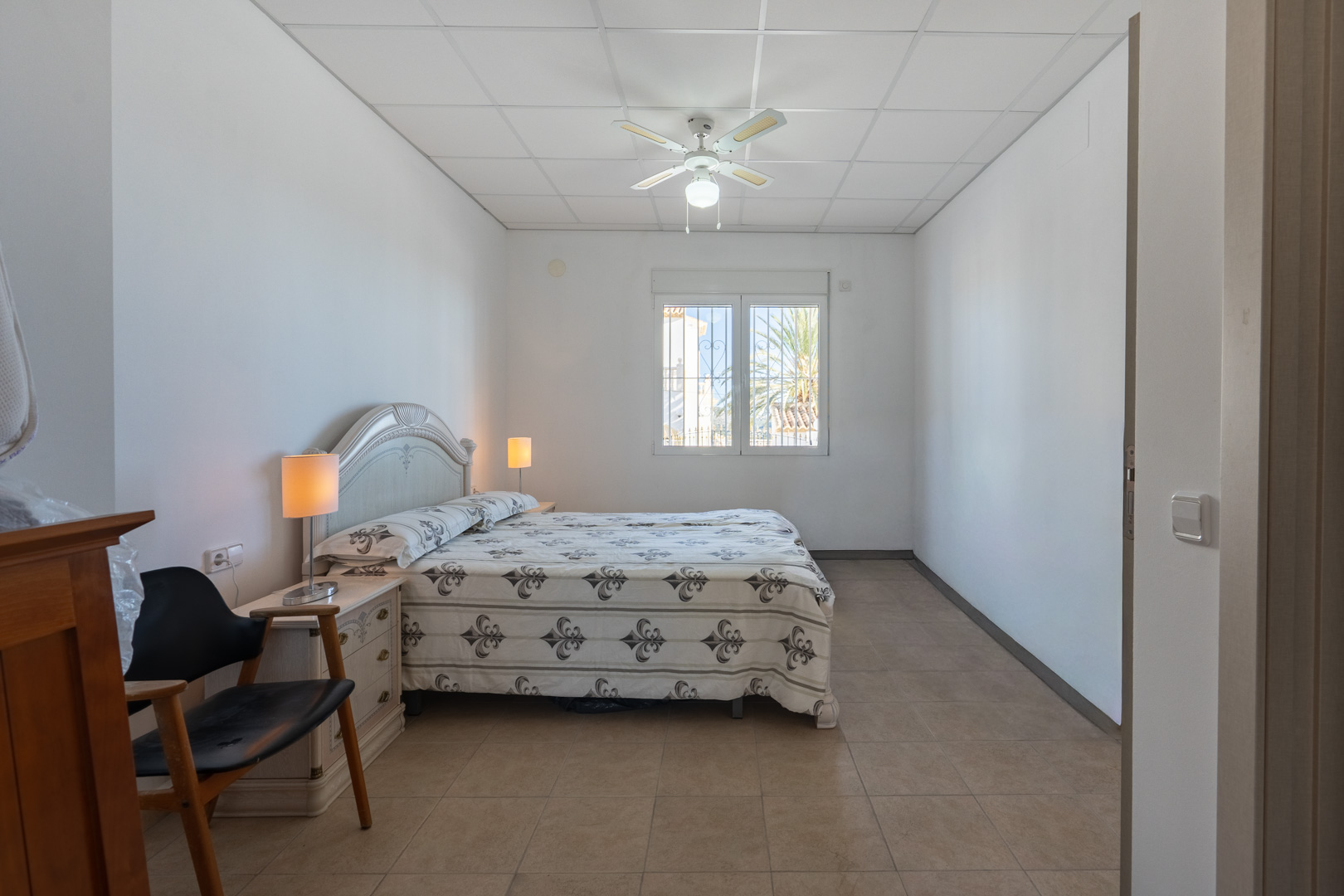 Helles Schlafzimmer mit Doppelbett, Deckenventilator und Fliesenboden. Ein Fenster sorgt für natürliches Licht und bietet Ausblick auf die mediterrane Flora.