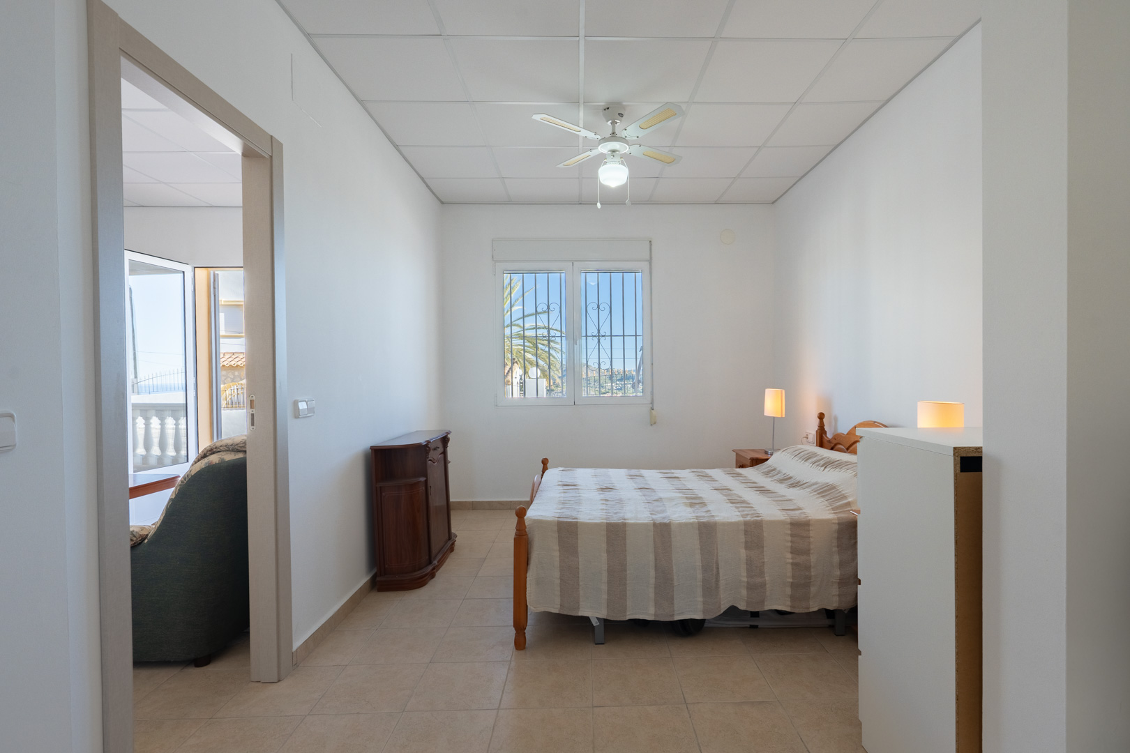 Helles Schlafzimmer mit Fliesenboden, Deckenventilator und Fenster mit Sicherheitsgittern. Der Raum bietet einen direkten Zugang zum Wohnbereich und Ausblick auf die Palmen.