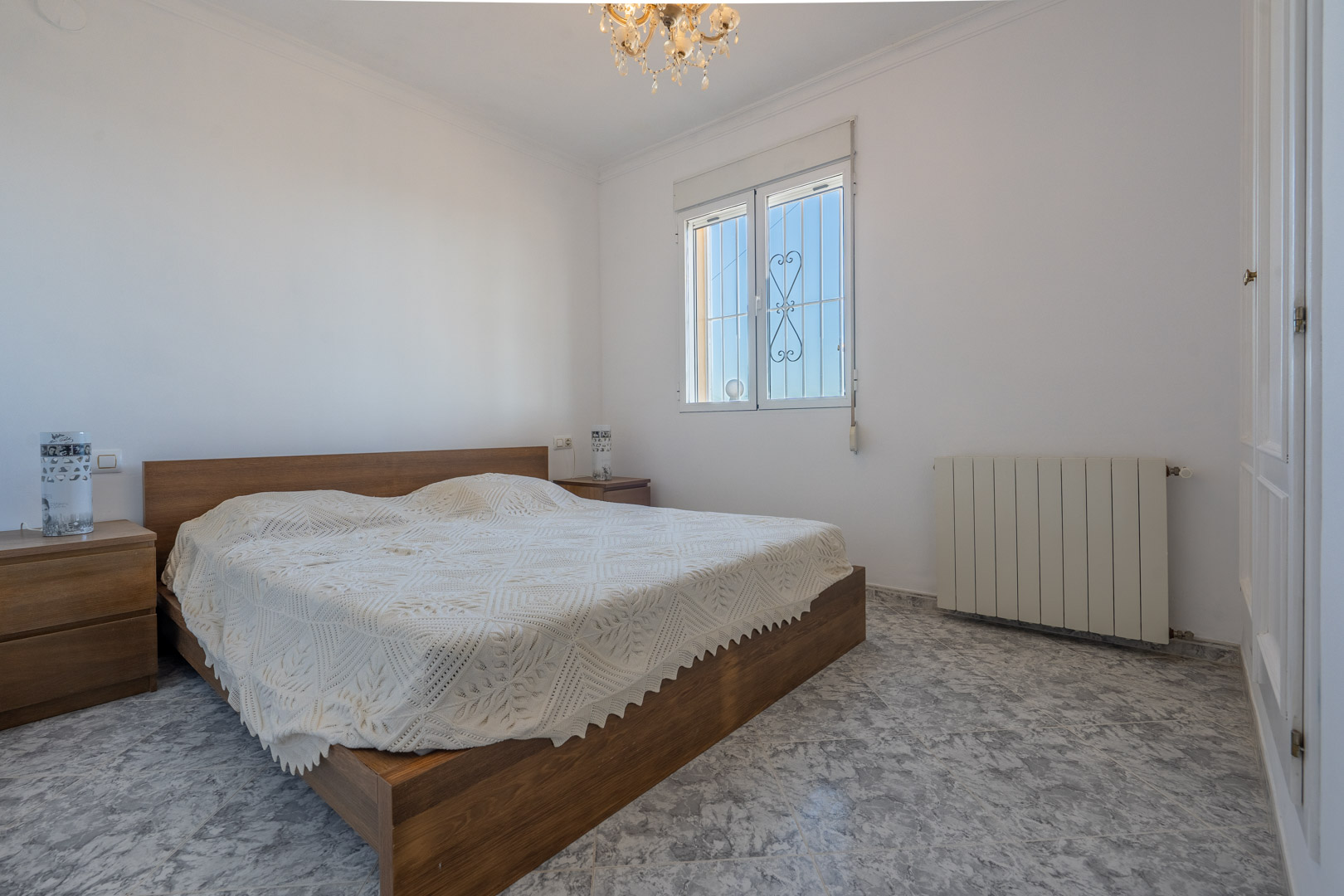 Helles Schlafzimmer mit Fliesenboden in Marmoroptik, Holzbett mit passenden Nachttischen, weißen Einbauschränken und Zentralheizung mit Heizkörper.