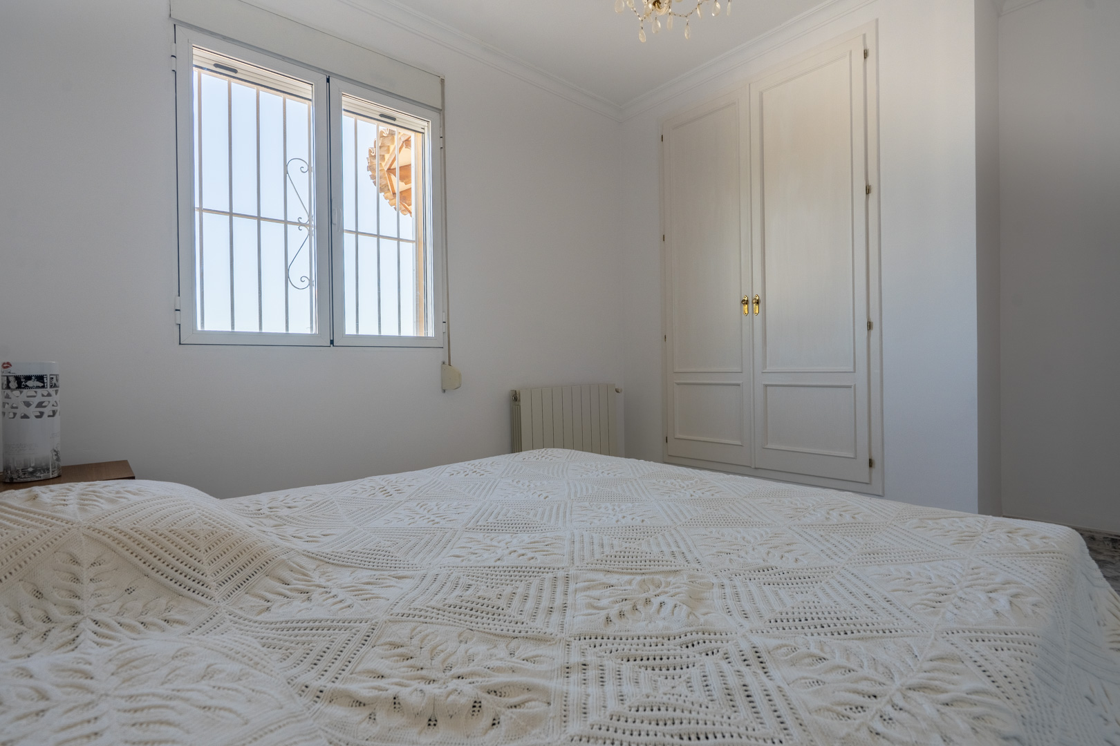 Helles Schlafzimmer mit weißen Wänden, einem Einbauschrank aus Holz und Fenster mit Sicherheitsgittern. Ausgestattet mit Heizkörper und natürlichem Lichteinfall.