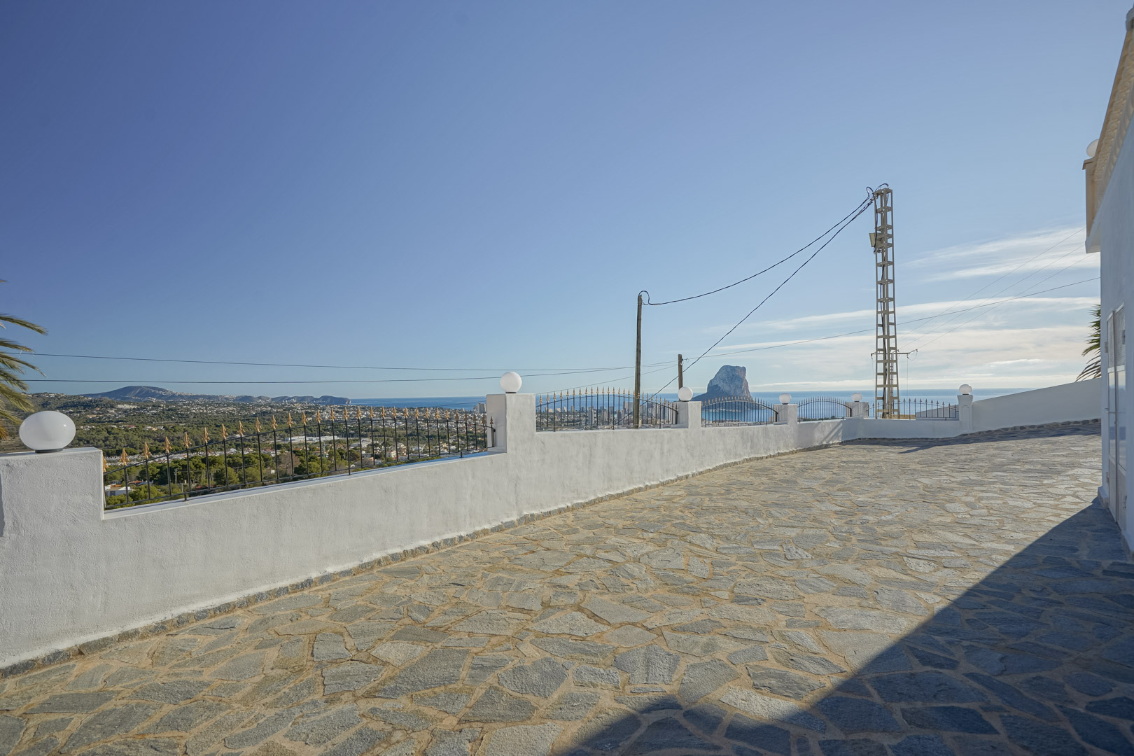 Mit Naturstein gepflasterte Terrasse mit Panoramablick auf das Mittelmeer und den Peñón de Ifach, eingefasst von einer weißen Mauer mit Schmiedeeisen.