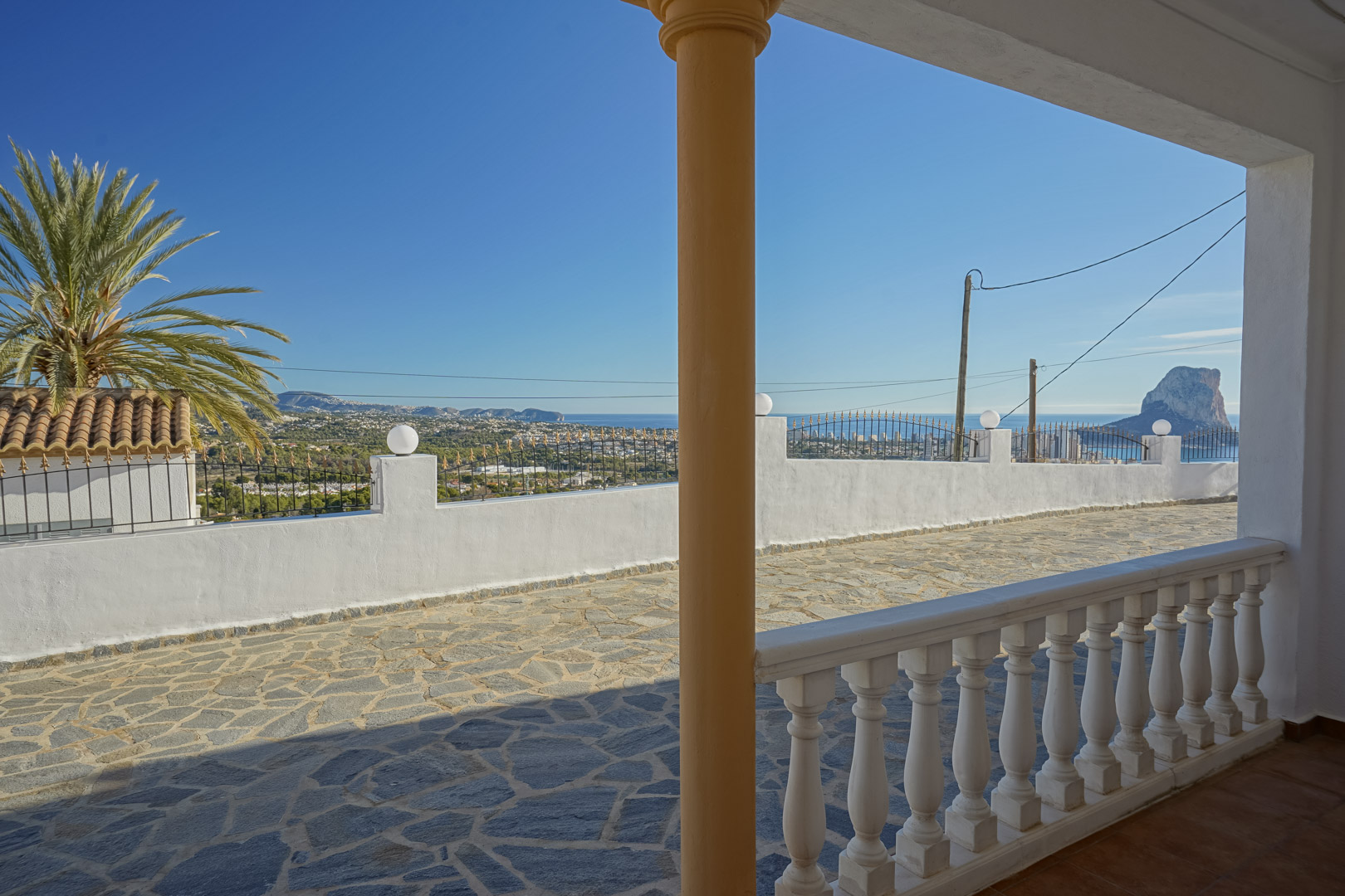Panoramablick von der überdachten Veranda mit weißer Balustrade auf die mit Naturstein gepflasterte Terrasse und den Peñón de Ifach in Calpe.