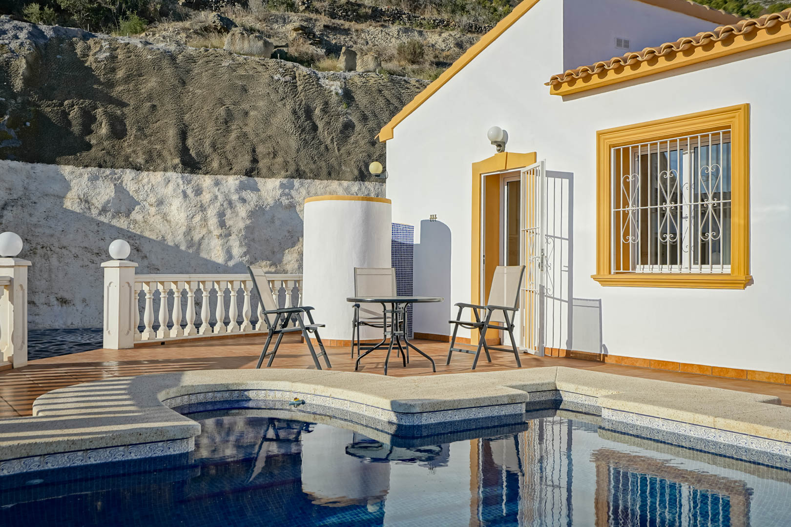 Poolterrasse mit Sitzgelegenheiten, zylindrischer Außendusche und weißer Fassade mit ockerfarbenen Fensterrahmen vor einer natürlichen Felswand.