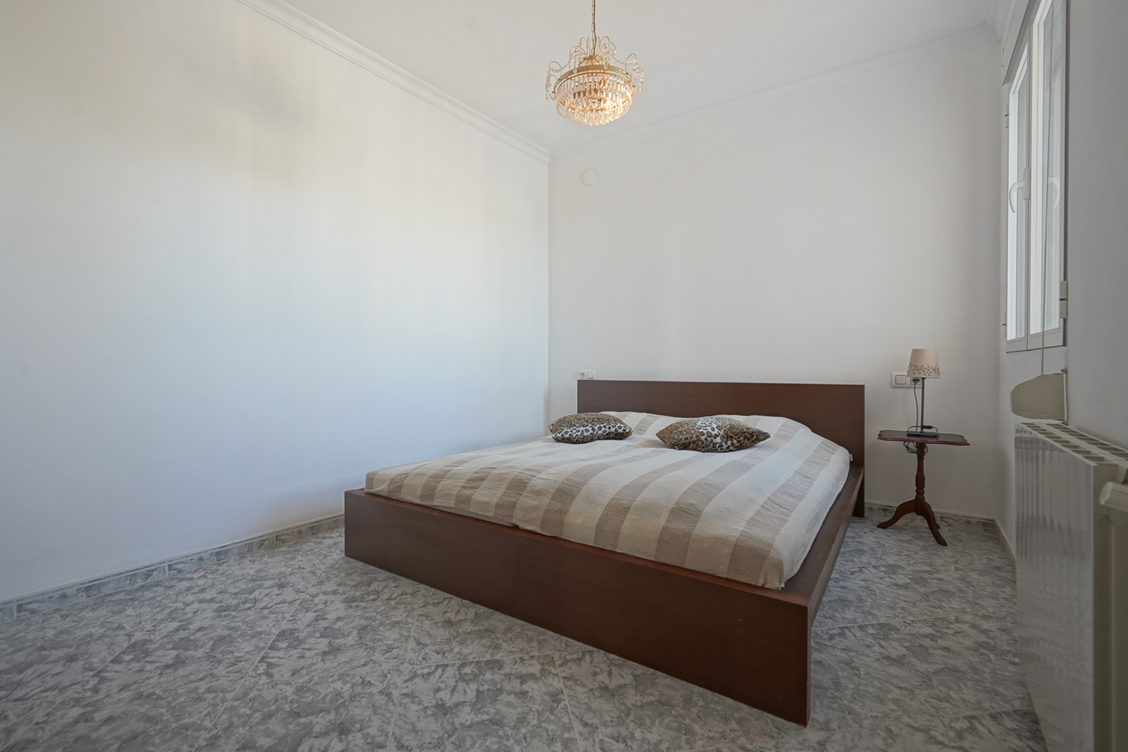Schlafzimmer mit dunklem Holzbettrahmen, marmorierten Fliesenböden, klassischem Kronleuchter und Heizkörper unter dem Seitenfenster.