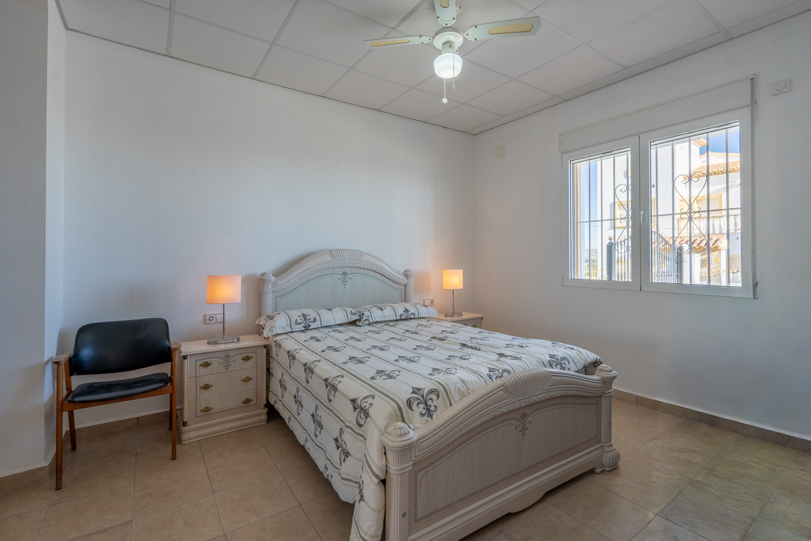 Schlafzimmer mit traditionellen Holzmöbeln, Fliesenboden, Deckenventilator und Fenster mit Sicherheitsgittern für natürliches Licht.