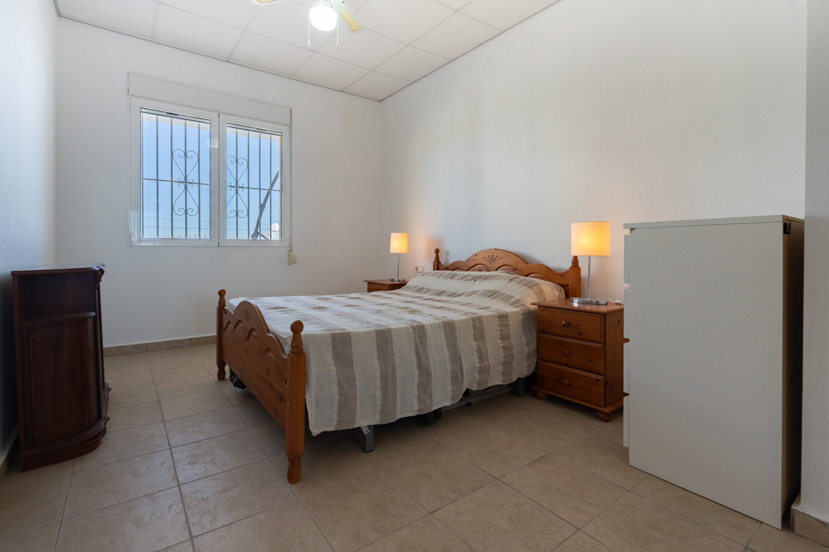 Schlafzimmer mit traditionellen Holzmöbeln, Fliesenboden und einem Fenster mit Sicherheitsgittern und Rollladen für natürliches Licht.