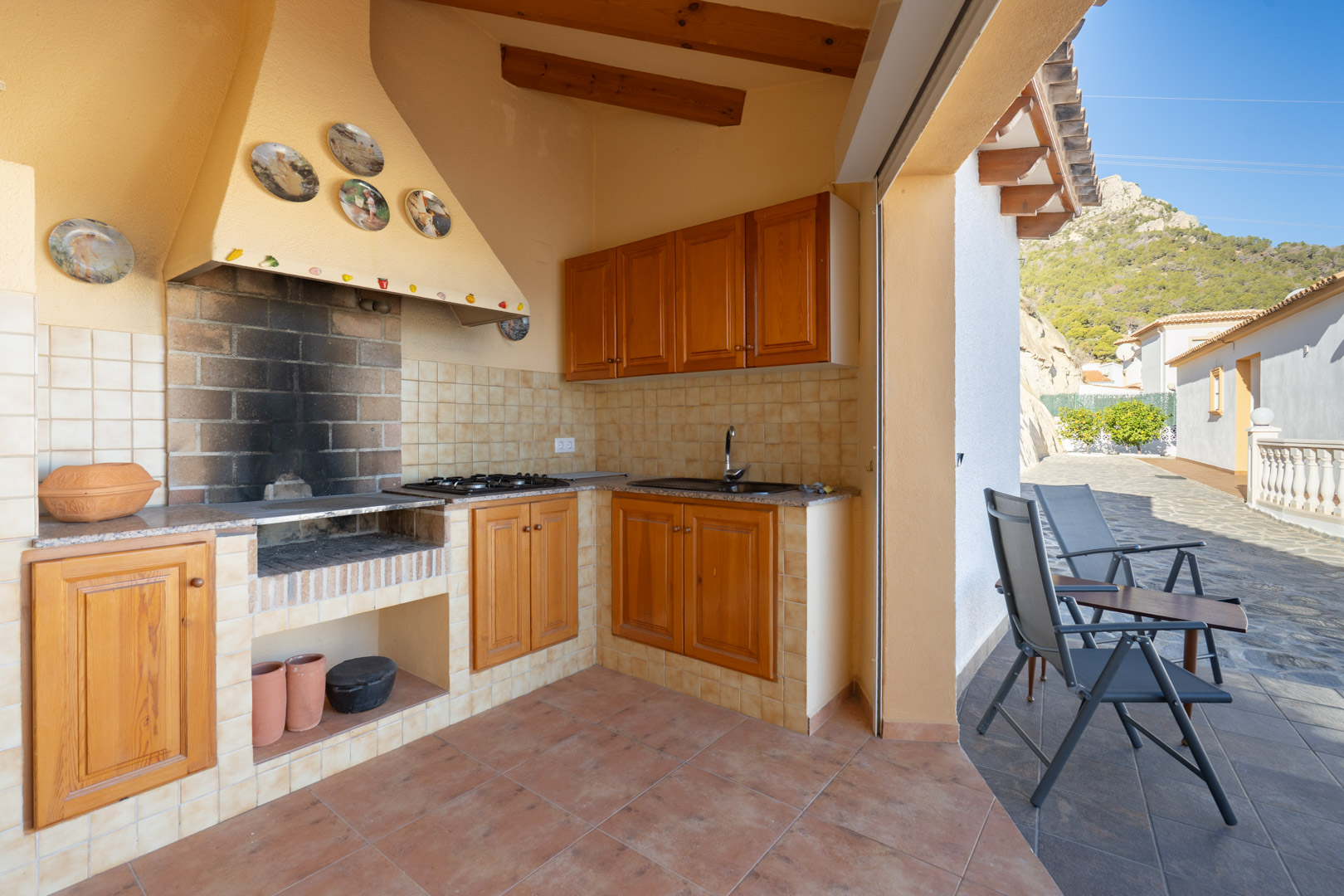 Überdachte Sommerküche mit gemauertem Grill, Holzschränken, Granitarbeitsplatte und Spüle, direkt an einer Terrasse mit Blick auf die Sierra de Oltà gelegen.