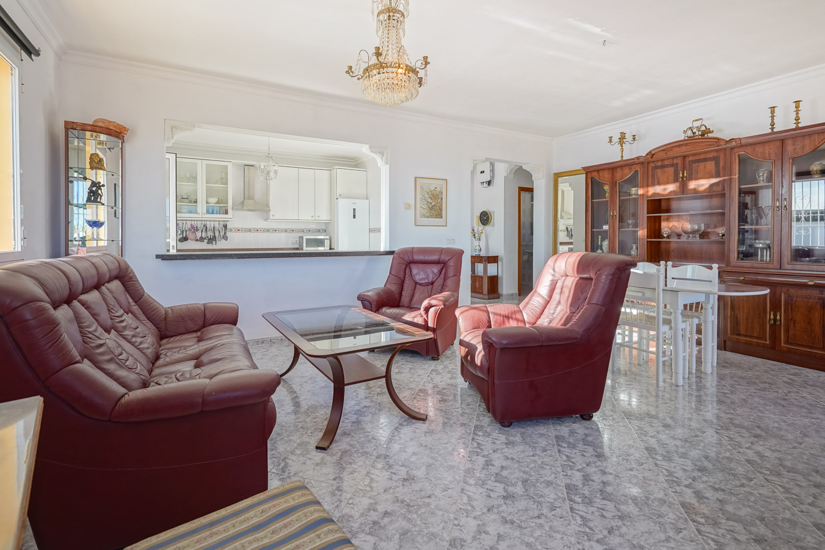 Wohn-Essbereich mit Marmorboden, burgunderroten Ledersofas und einer halb offenen Küche. Ein klassischer Holzschrank und ein Kristallleuchter prägen das Interieur.