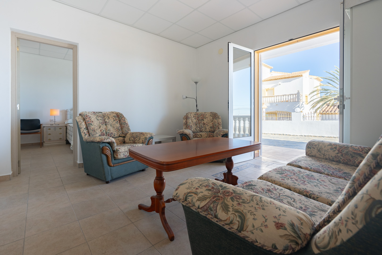Wohnbereich der Einliegerwohnung mit floralen Polstermöbeln, Holztisch und Zugang zur Terrasse. Der Raum verfügt über Fliesenboden und Blick in ein Schlafzimmer.