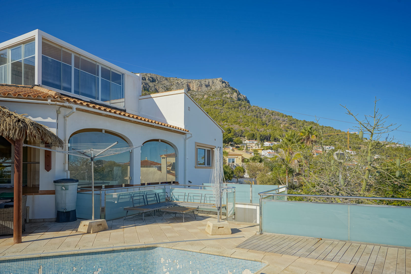 Außenansicht der Villa mit Fliesenterrasse, Glasgeländern und Blick auf die Sierra de Oltamar. Mit Rundbogenfenstern und verglastem Obergeschoss neben dem Poolbereich.