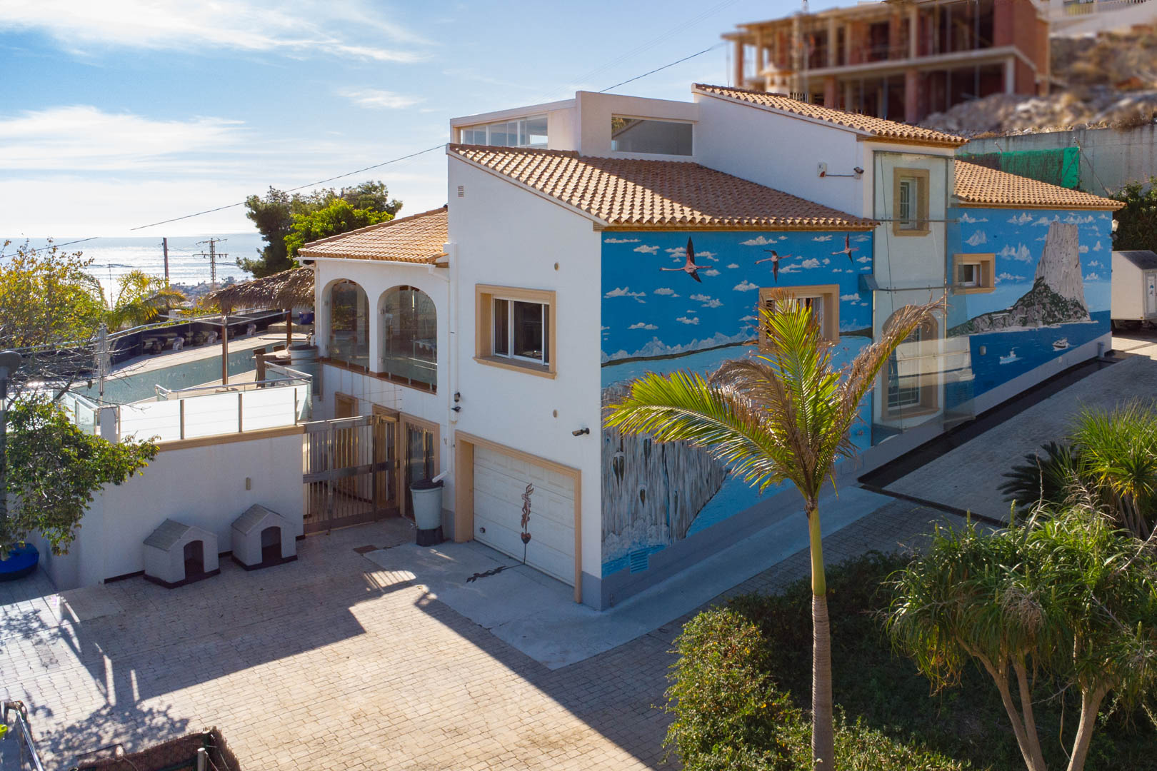 Außenansicht der Villa mit markantem Wandgemälde des Peñón de Ifach, Garagenzufahrt und verglasten Terrassenbereichen vor Küstenkulisse.