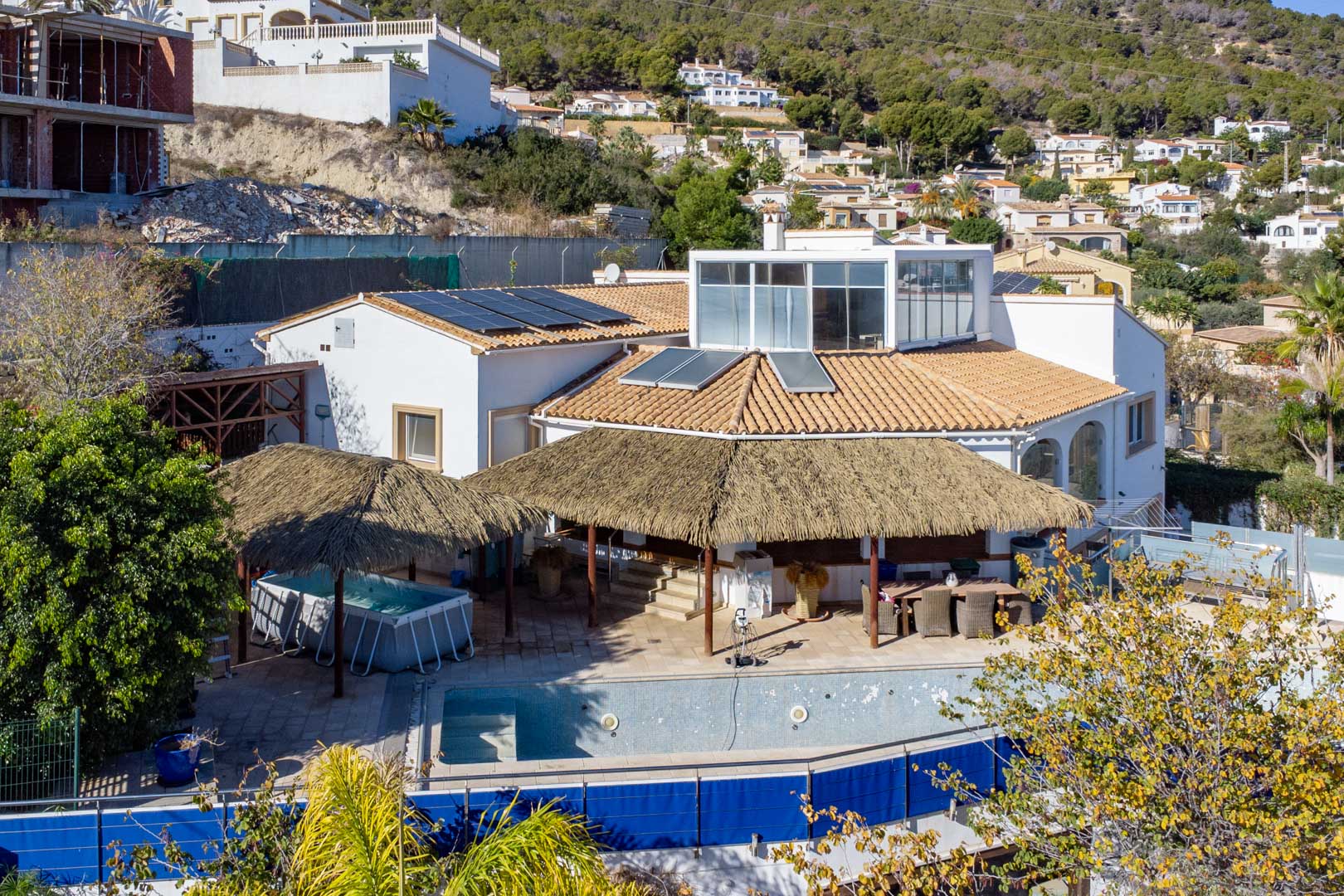Außenansicht der Villa mit Solaranlagen, Terrassen mit Rieddach-Pergolen, Poolbereich und einer verglasten Dachterrasse in Hanglage von Calpe.