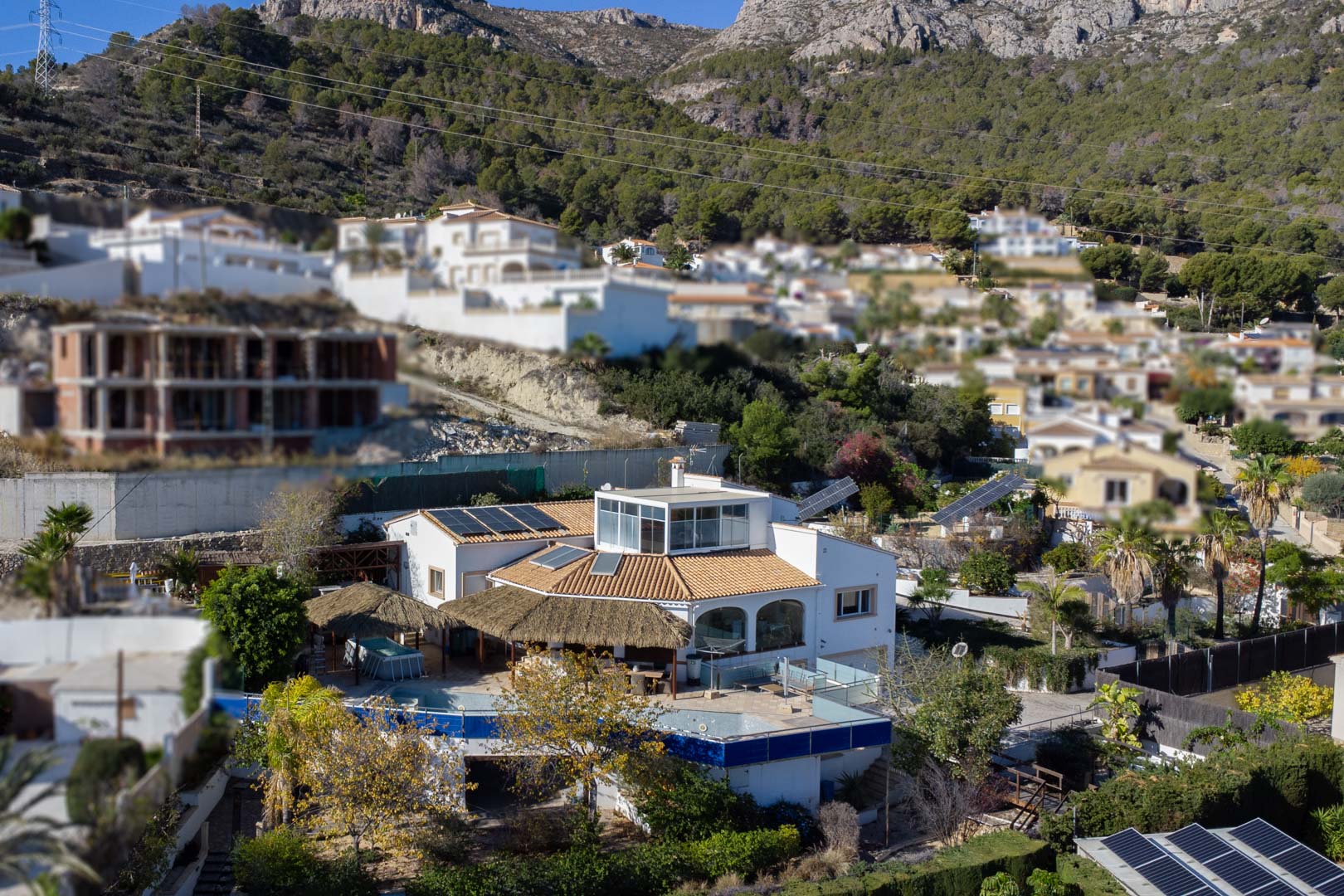 Außenansicht der Villa mit Solaranlagen, verglaster Oberterrasse und Poolbereich mit Reetdach-Pergolen vor der Kulisse der Sierra de Oltamar.