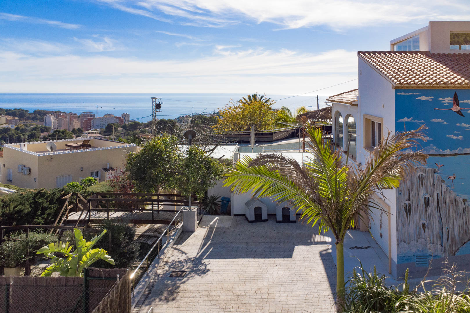 Außenansicht mit gepflasterter Einfahrt, Flamingo-Wandbild an der Fassade und Panoramablick auf das Mittelmeer und die Skyline von Calpe.