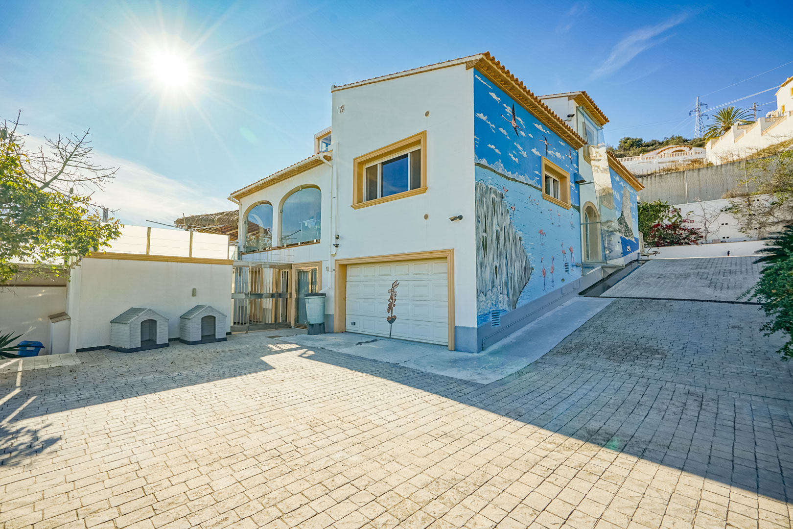 Außenansicht mit großzügiger gepflasterter Einfahrt, integrierter Garage und einem markanten Wandgemälde mit Küstenmotiven an der Seitenfassade.