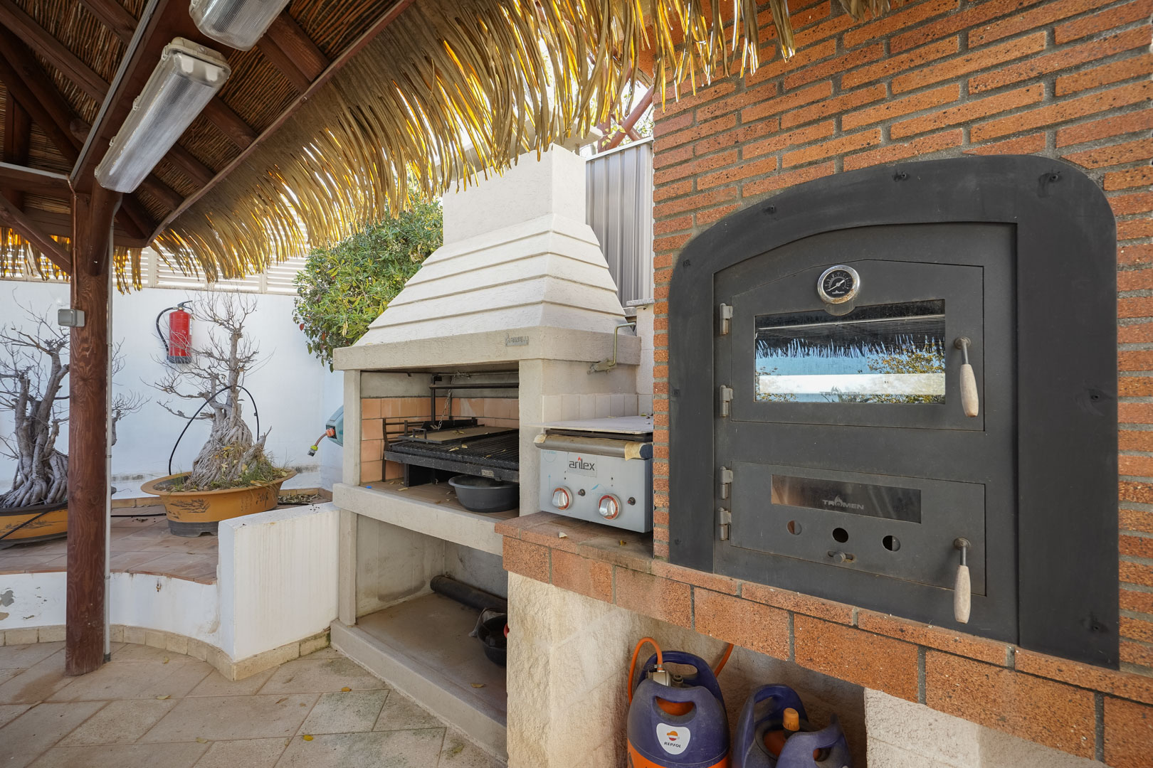 Außenkochbereich mit eingebautem Tromen-Ofen, Grillstation und Gasgrill unter einer rustikalen Holzpergola mit natürlicher Reetbedeckung.
